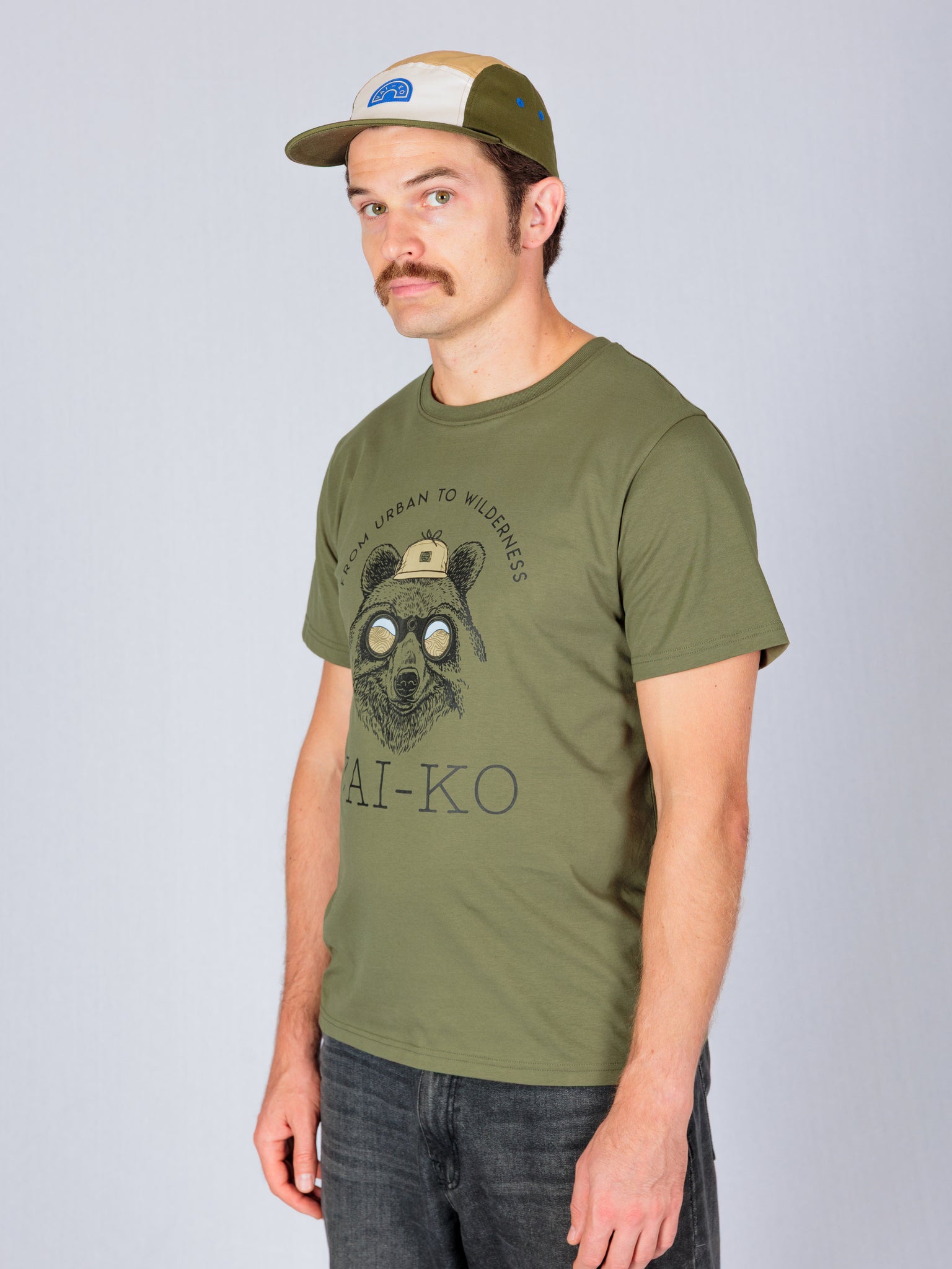 Man in a green graphic t-shirt and cap standing against a plain light background, looking at the camera.