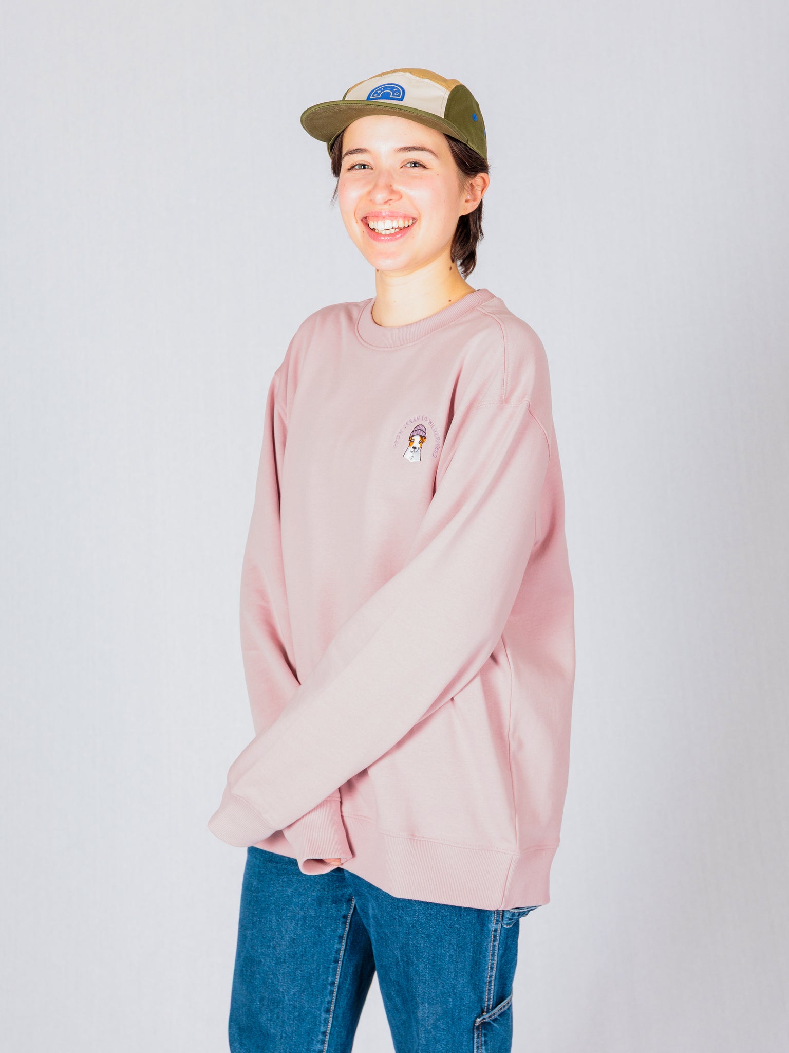 Smiling person in a pink sweatshirt, blue jeans, and a beige cap stands against a plain light background.