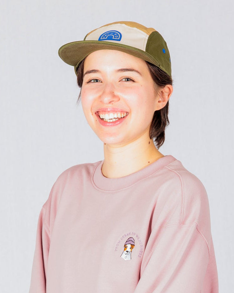 Smiling person in a pink sweatshirt, blue jeans, and a beige cap stands against a plain light background.