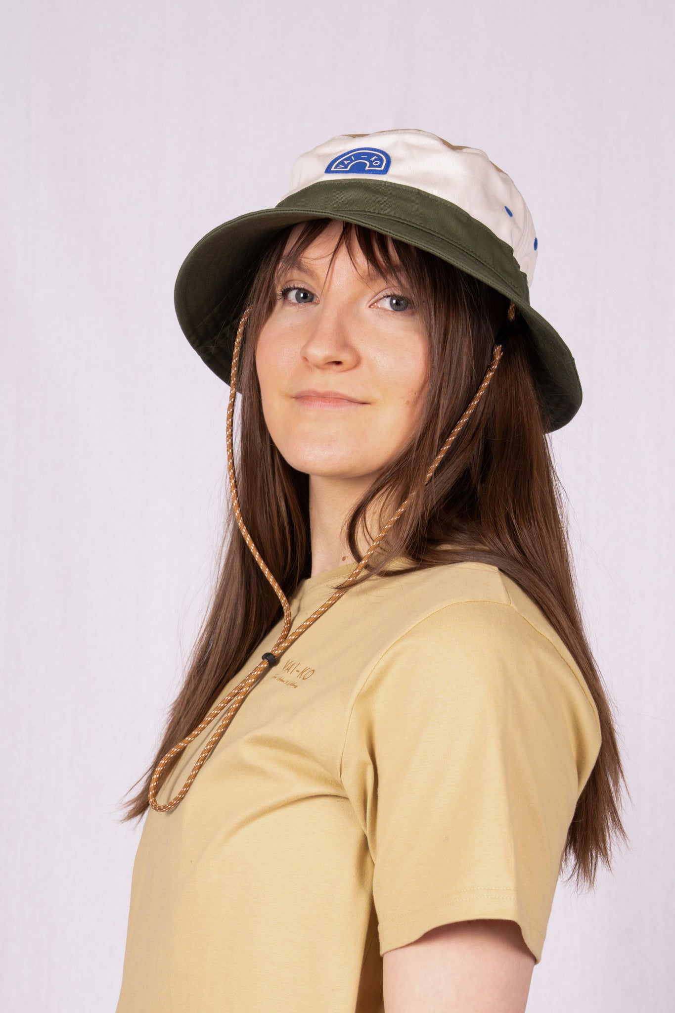 Woman in beige shirt and bucket hat with chin cord, standing against a light background, looking at camera.