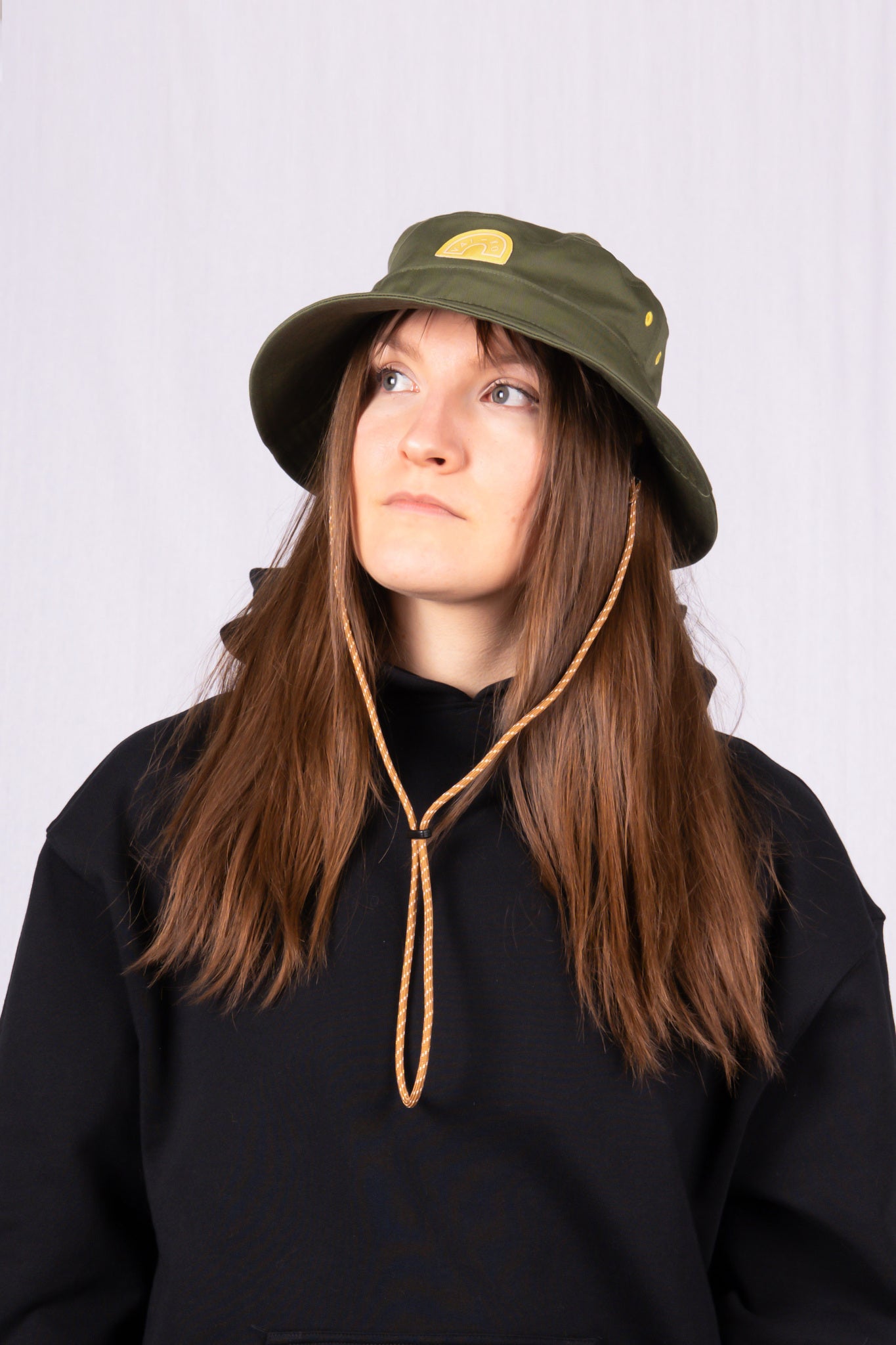 Woman in a green bucket hat and black hoodie looking up, with long brown hair and a neutral expression.