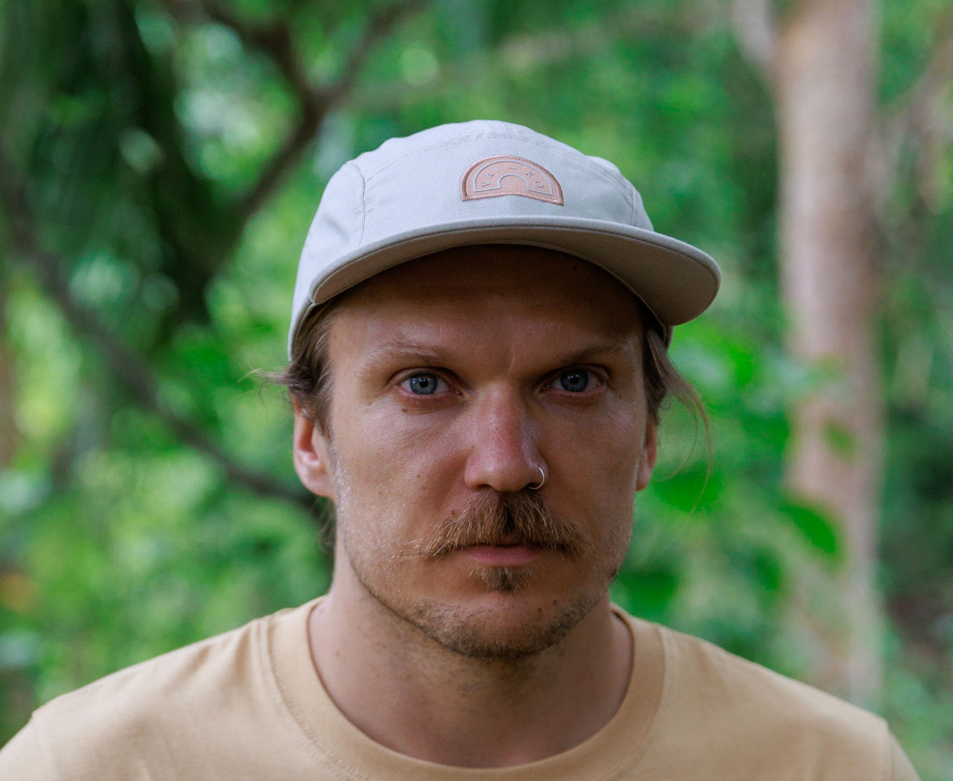 A man with a mustache and cap stares at the camera, with greenery blurred in the background.
