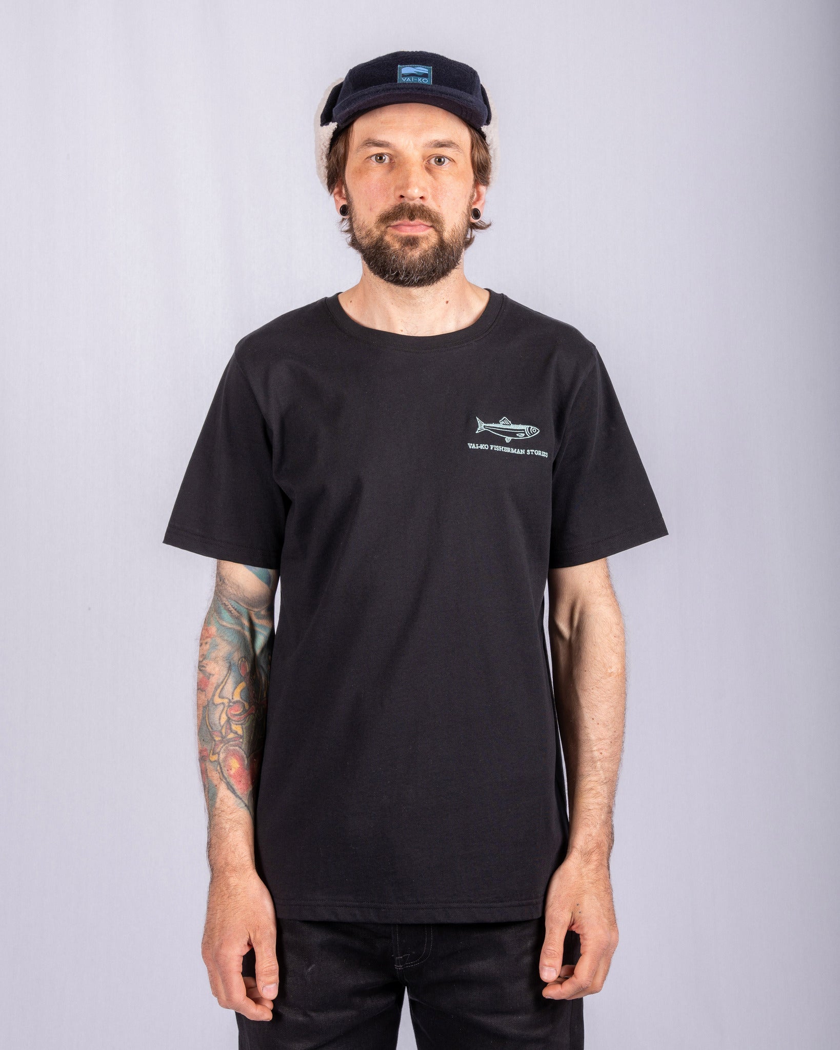 Man in black t-shirt and cap stands in front of a plain light background, showing tattooed right arm.