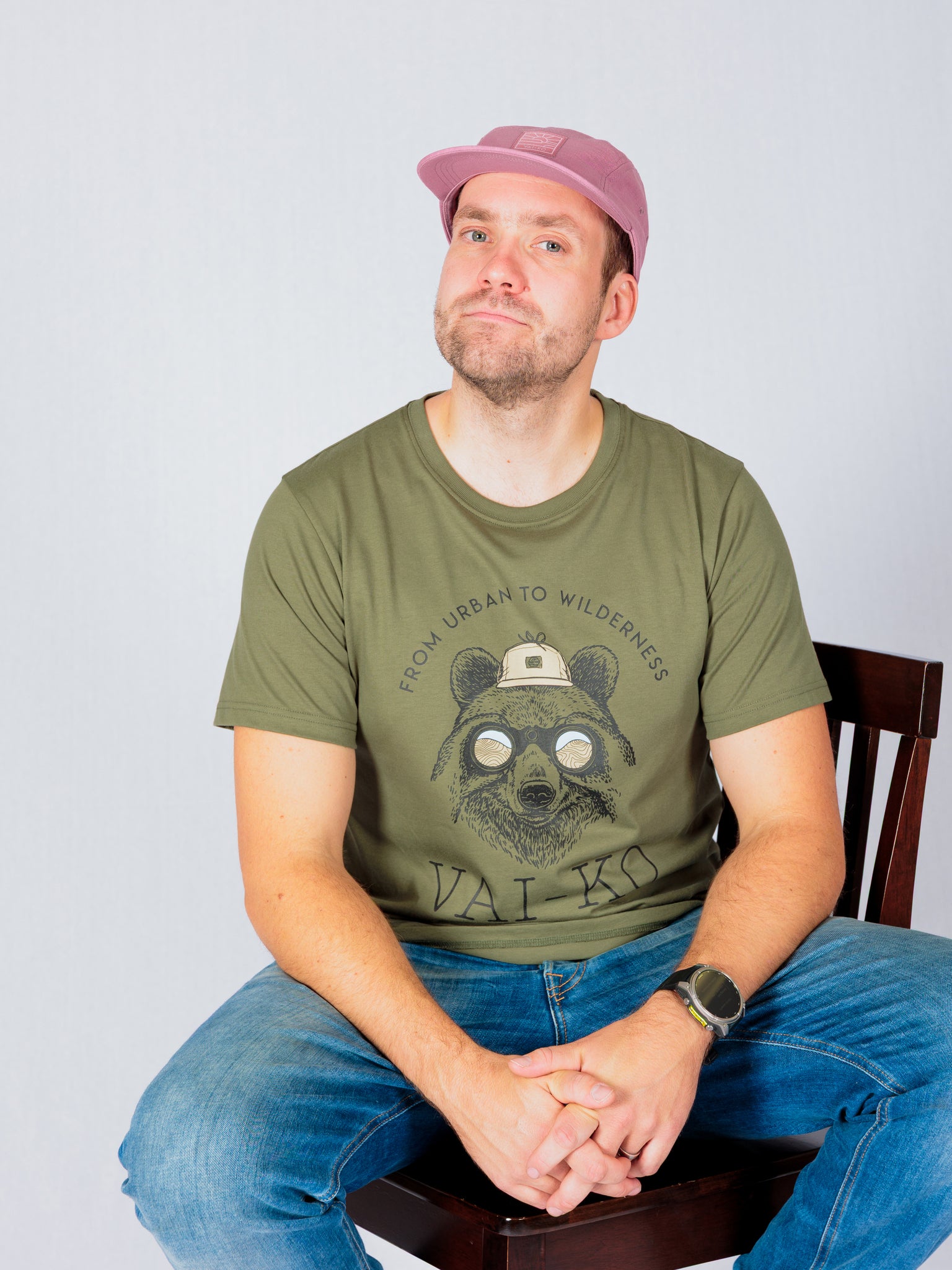 Man in pink cap and green t-shirt with bear graphic sits on a chair, smiling slightly at the camera.