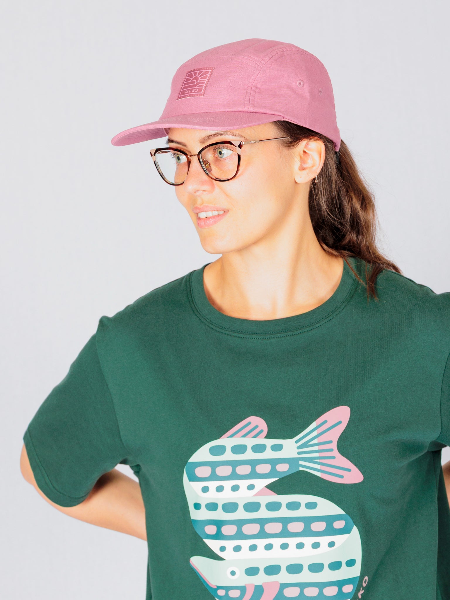 Woman in glasses wearing a pink cap and a green shirt with a colorful fish graphic, looking to the side.