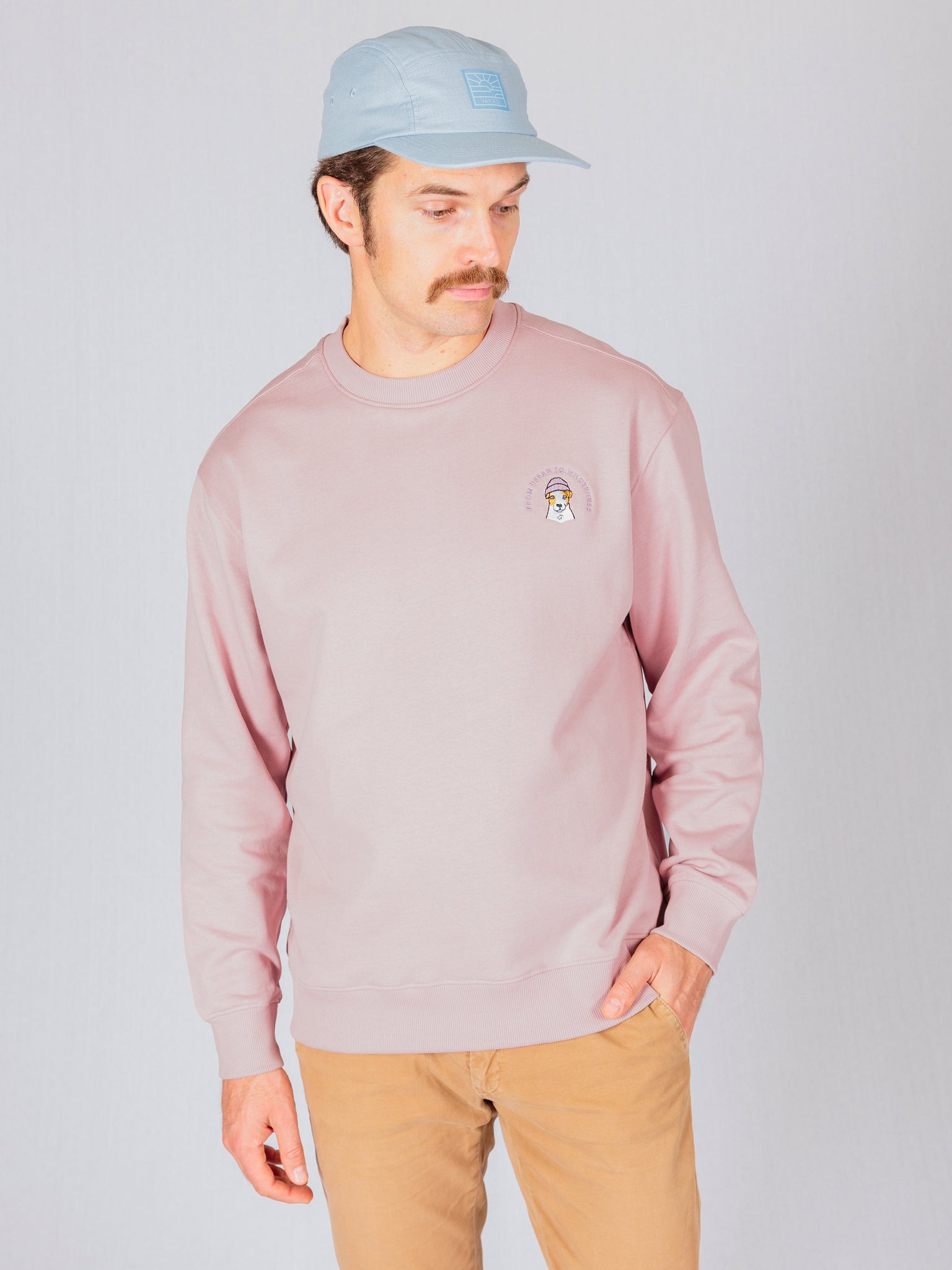 Man wearing a light pink sweatshirt, tan pants, and a light blue cap, looking down with one hand in his pocket.