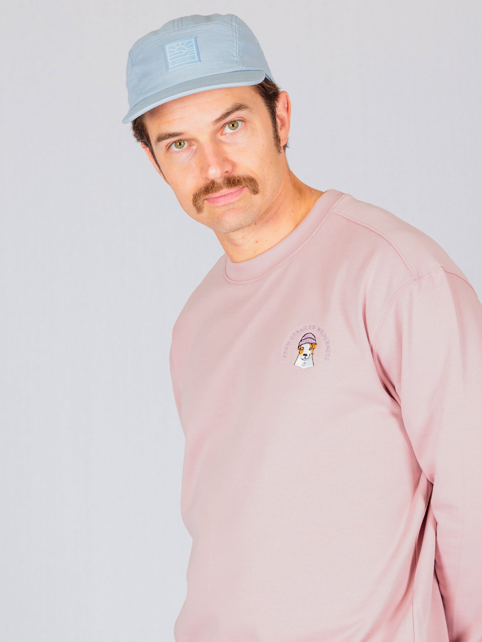 Man with a mustache wearing a light blue cap and pink sweatshirt, standing against a plain background.