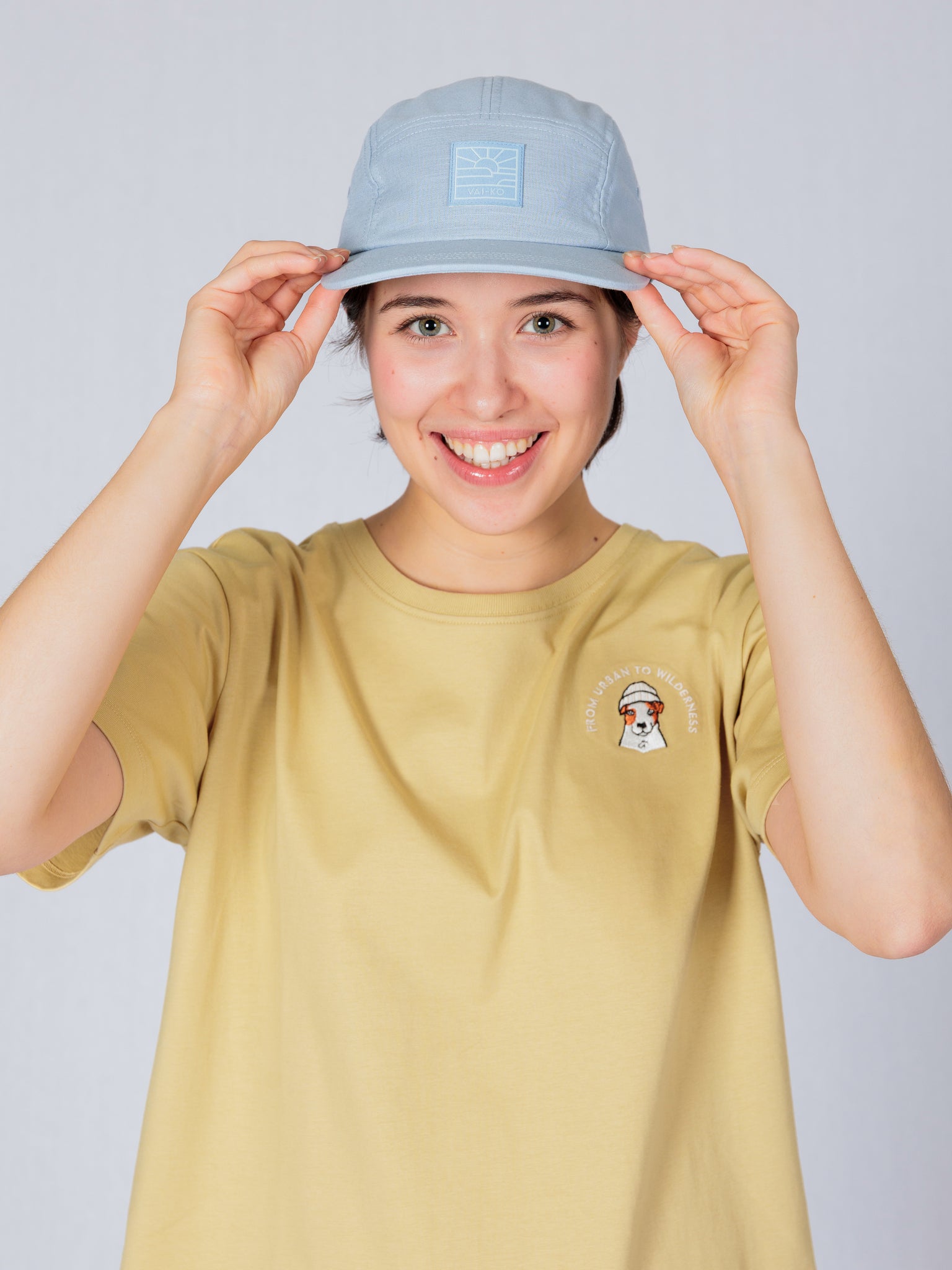 Smiling person in a beige t-shirt and light blue cap, holding the brim of the cap with both hands.