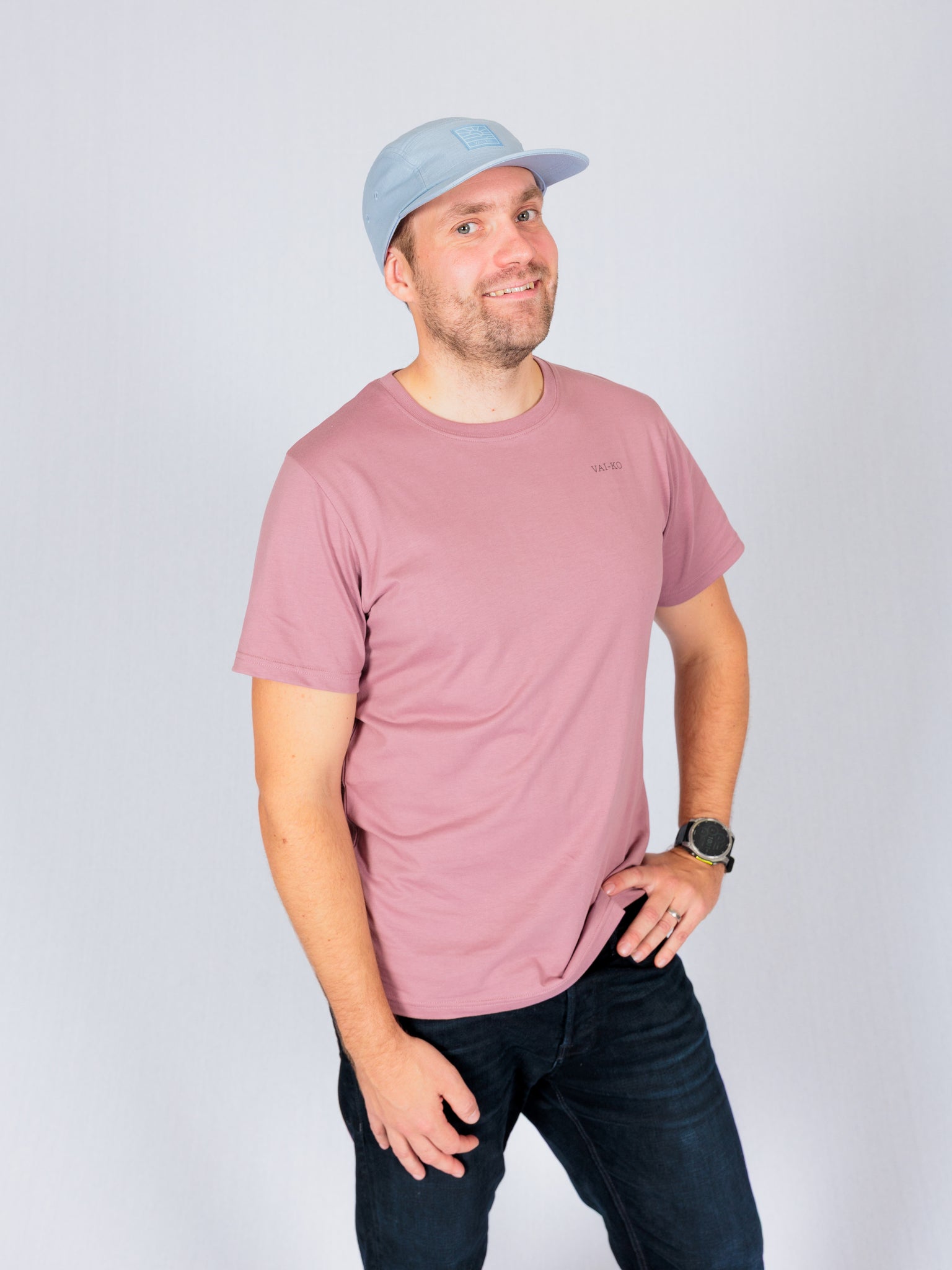 Man in a pink t-shirt and light blue cap smiling, standing with one hand on hip against a plain background.