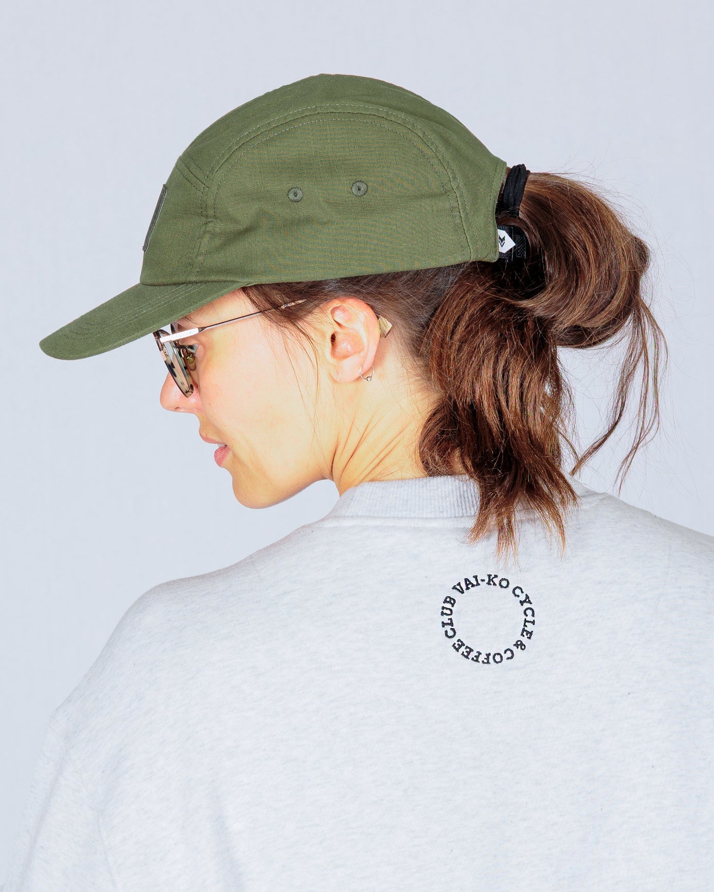 Woman in green cap, sunglasses, and gray shirt with black circular text on the back, facing away.