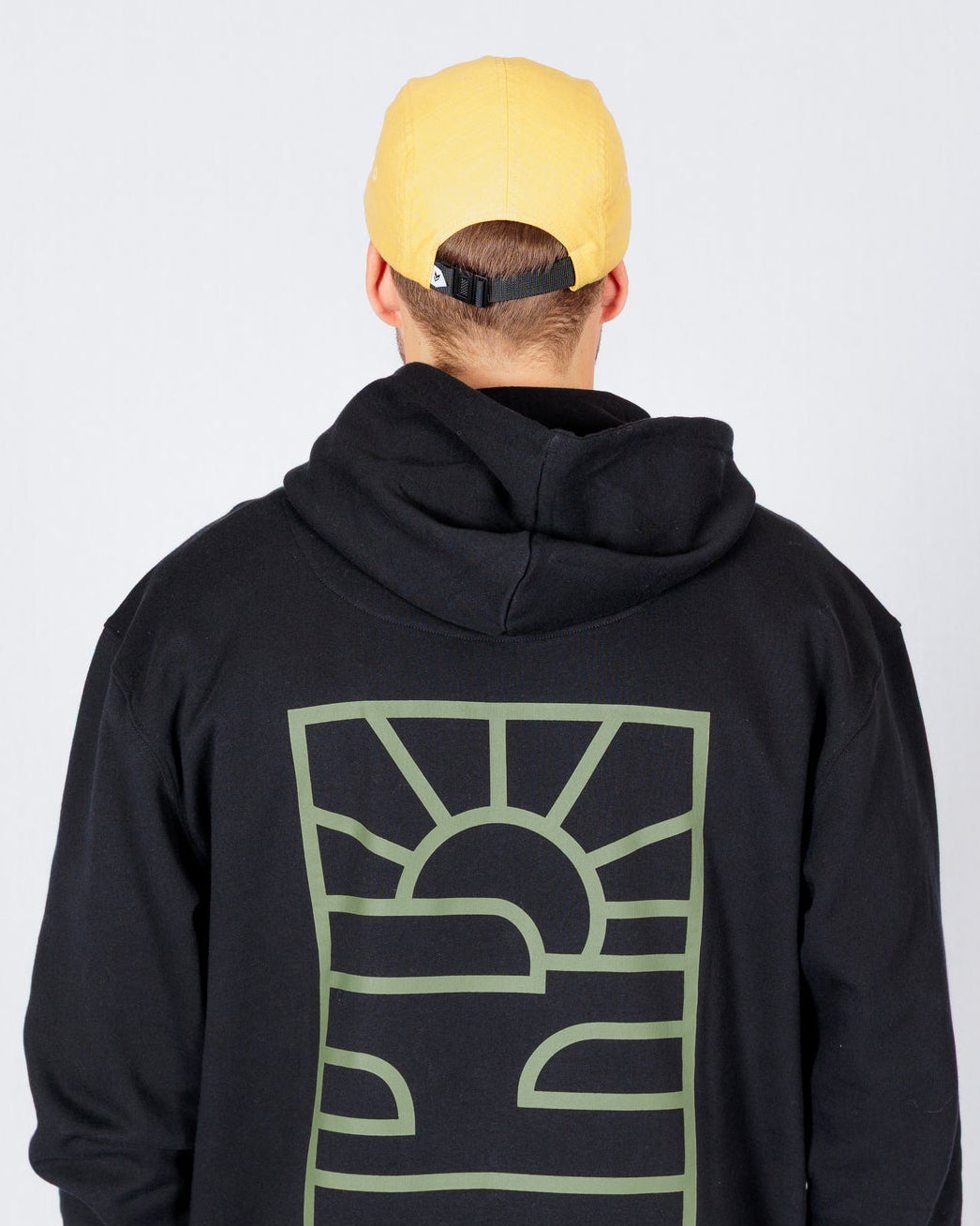 Person in a yellow cap and black hoodie with a graphic sun and landscape design on the back stands facing away.