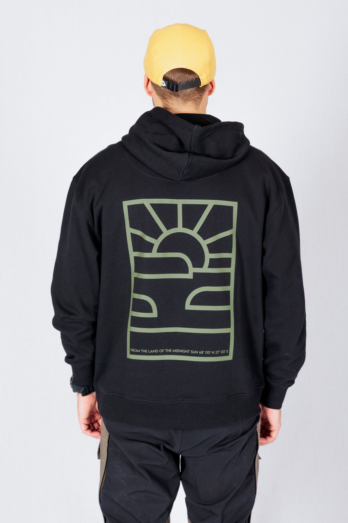 Person in a yellow cap and black hoodie with a graphic sun and landscape design on the back stands facing away.