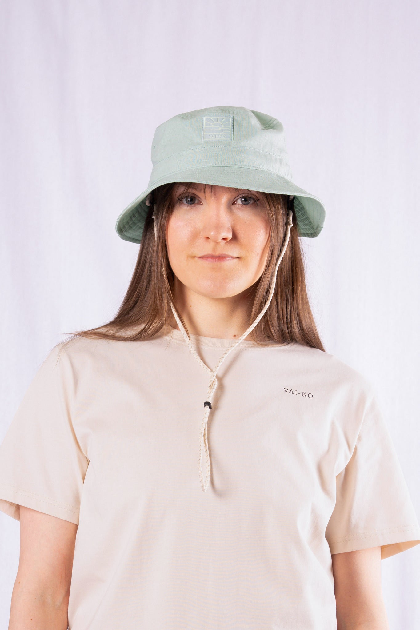 A person wearing a light green bucket hat and a beige T-shirt with the text VAI-KO on it.