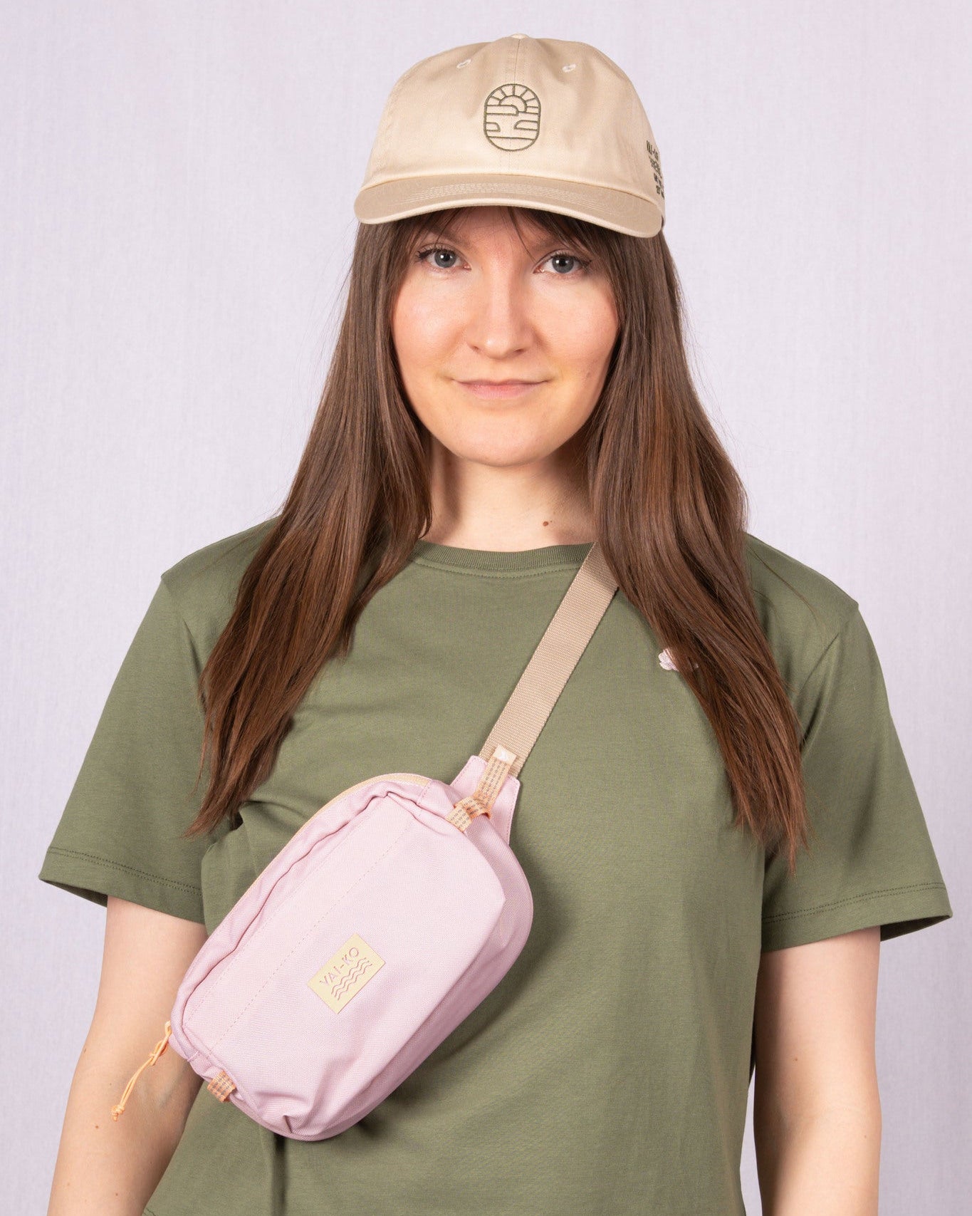 Woman in a green t-shirt, beige cap, and pink crossbody bag standing against a light gray background.