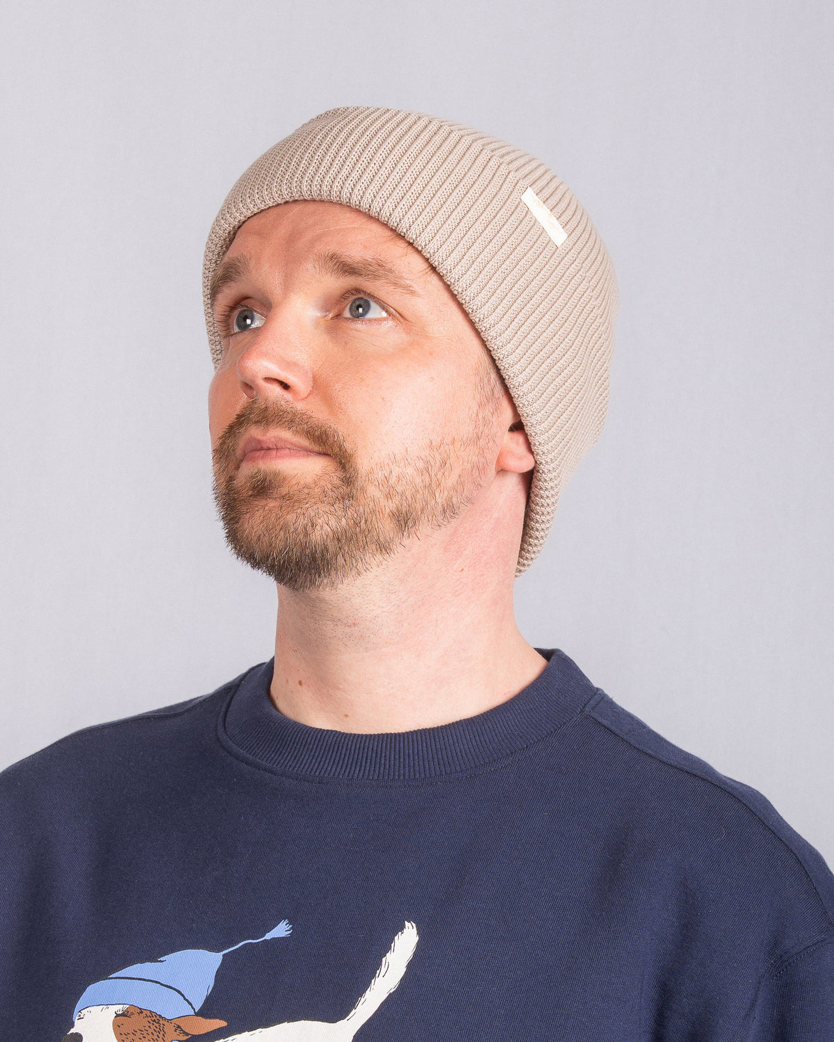 Man with a beard wearing a beige knit beanie and navy sweater, looking upwards against a plain background.