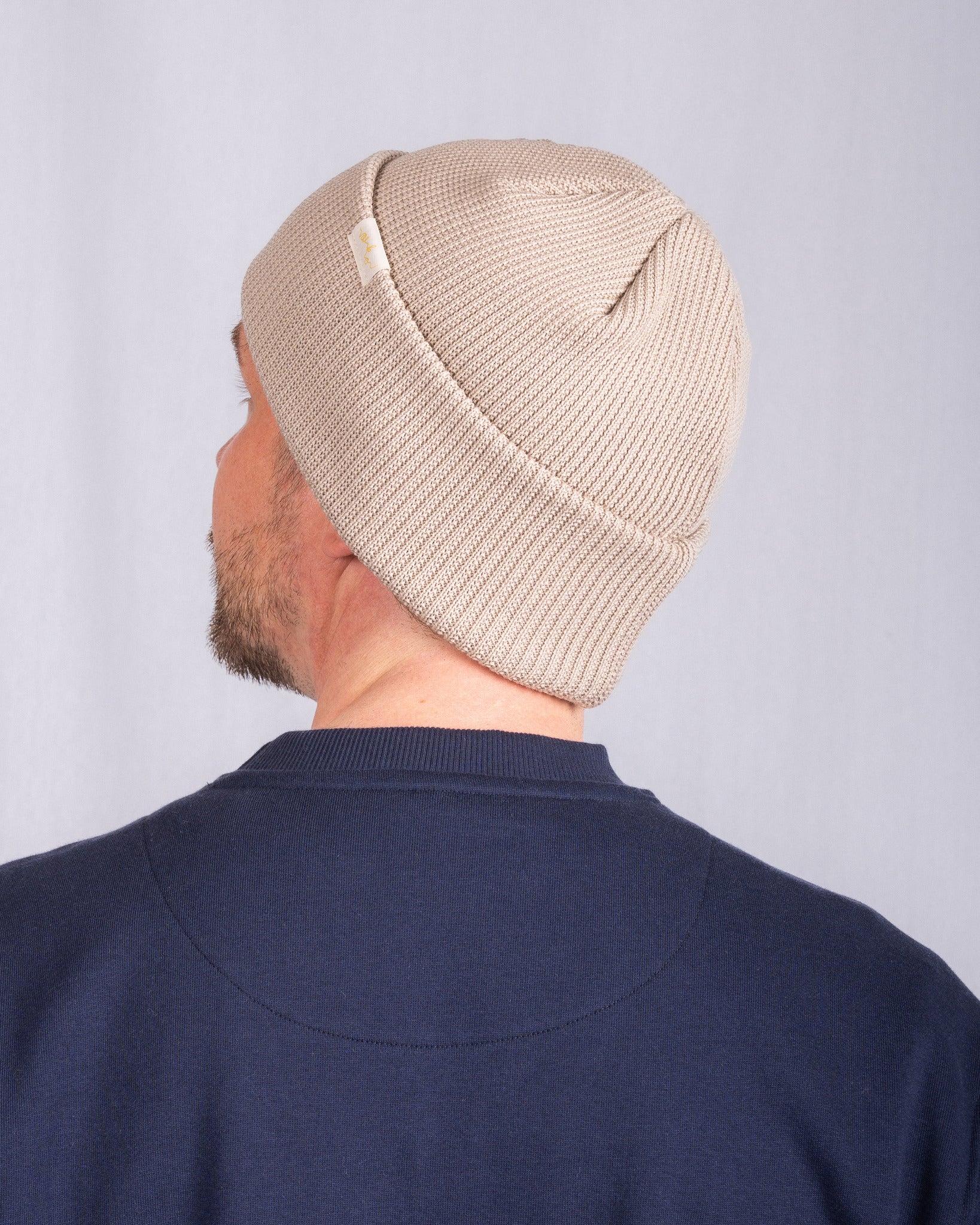 A person wearing a beige knit beanie and a navy blue shirt, shown from the back against a plain background.