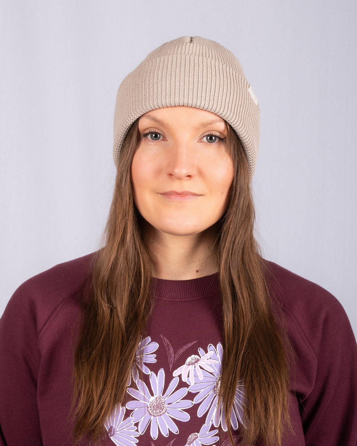 Woman in beige knit beanie and maroon sweater with purple daisies, facing forward against plain background.