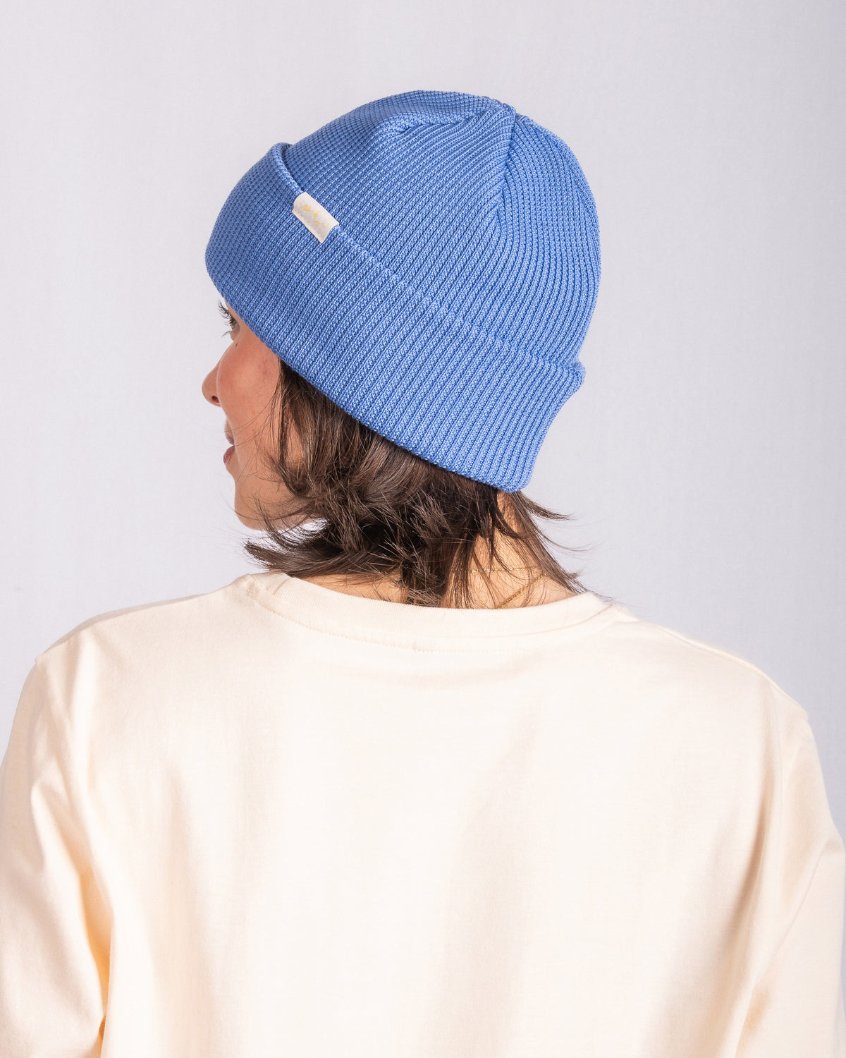 Person wearing a blue knit beanie and cream shirt, facing away from the camera against a plain background.