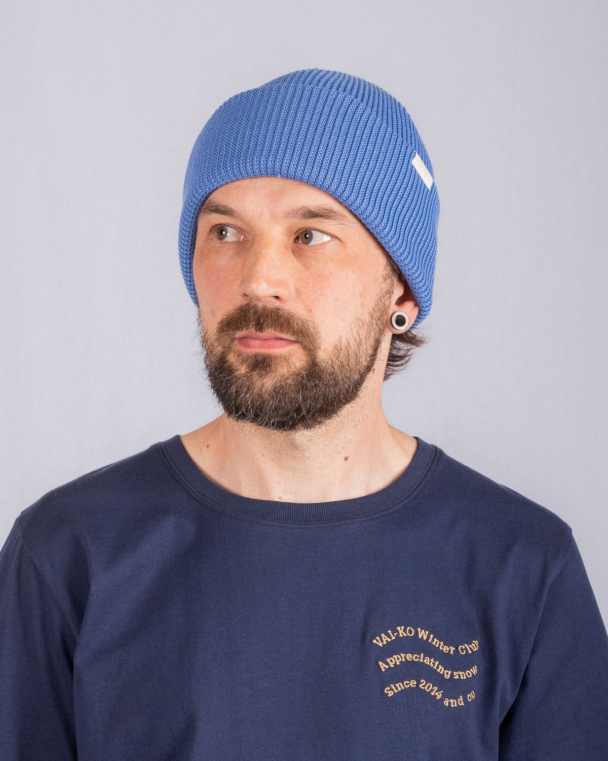 Bearded man in a blue beanie and navy shirt looks to the left, with a neutral expression.