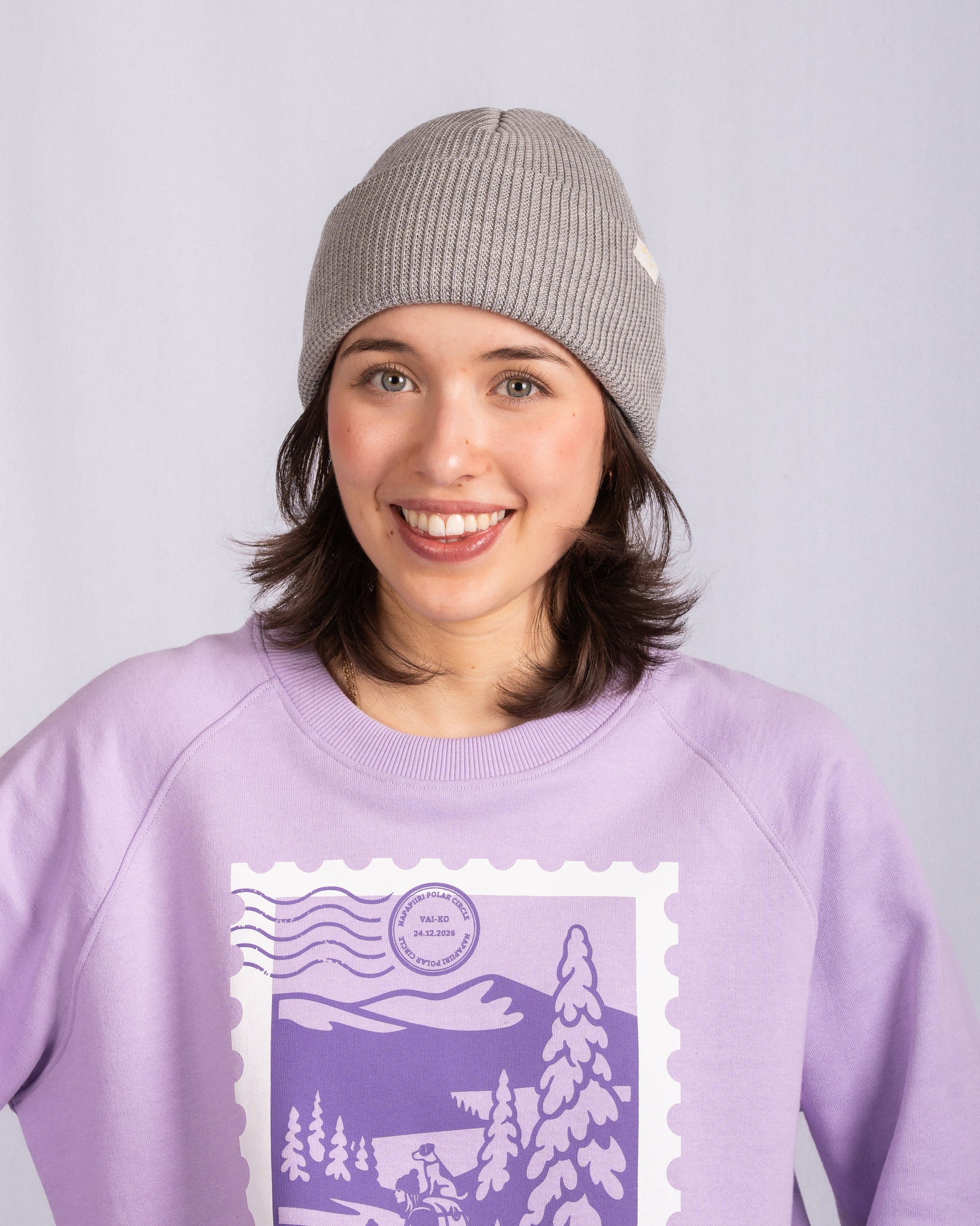 Smiling person in a gray beanie and purple sweatshirt with a winter scene design on the front.