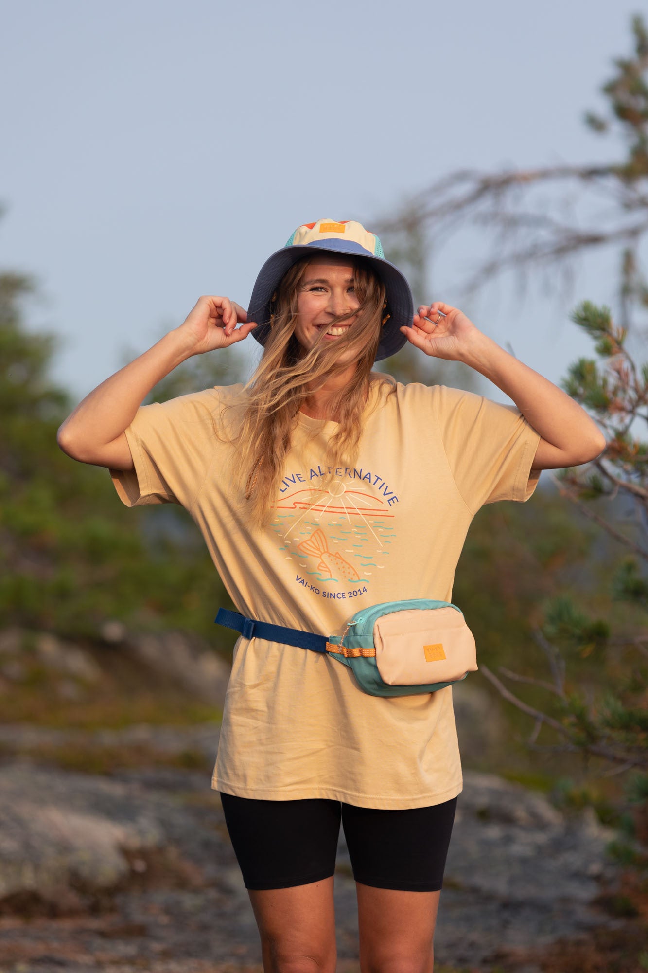 Smiling woman in a hat and t-shirt stands outdoors, adjusting her hat, with trees in the background.