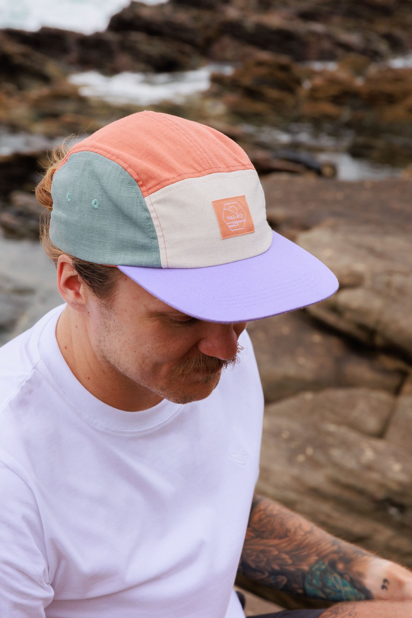 Man in a white shirt wearing a multicolored cap sits outdoors on rocks near the water.