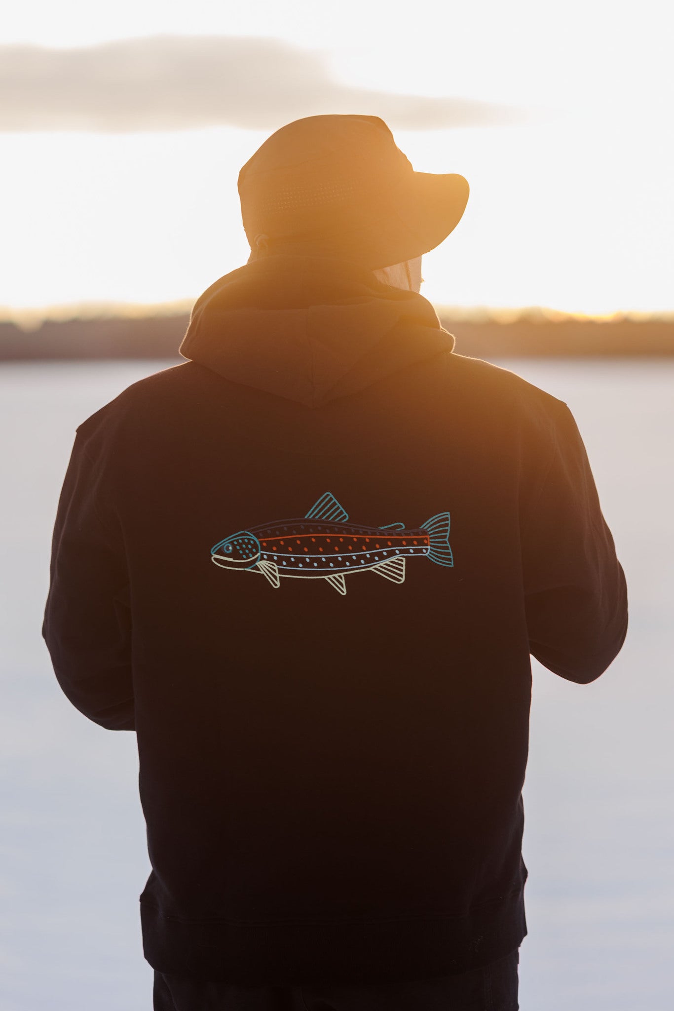 Person in a hat and hoodie with a colorful fish graphic on the back, standing outdoors at sunset.
