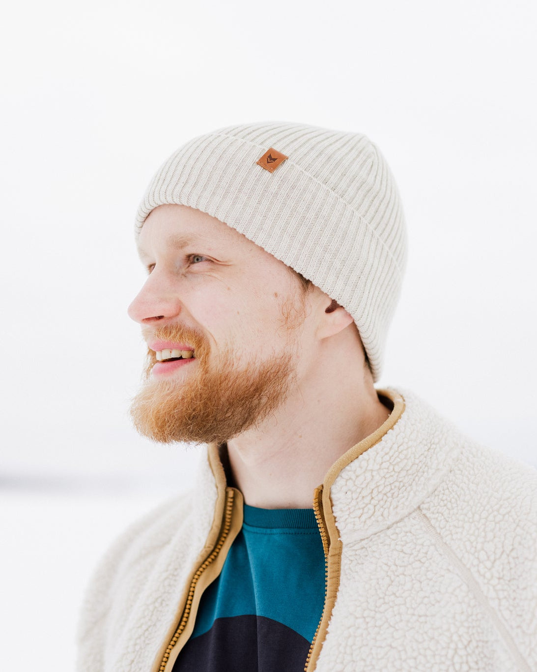 Bearded man in a knit hat and fleece jacket smiling outdoors in a snowy, bright setting.