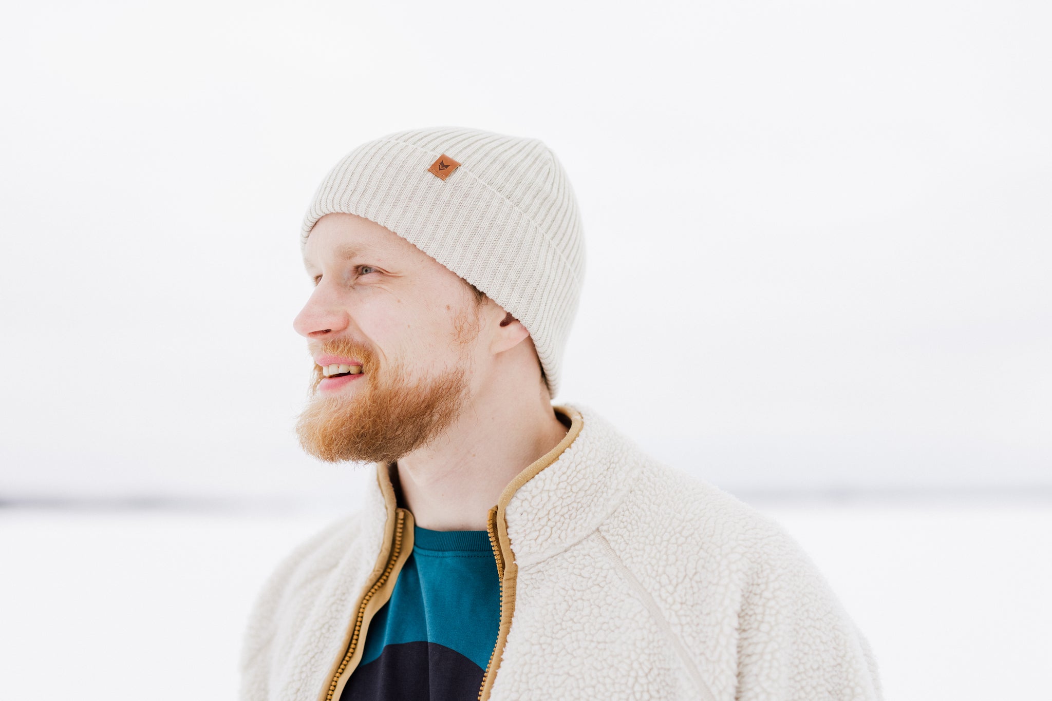 Bearded man in a knit hat and fleece jacket smiling outdoors in a snowy, bright setting.