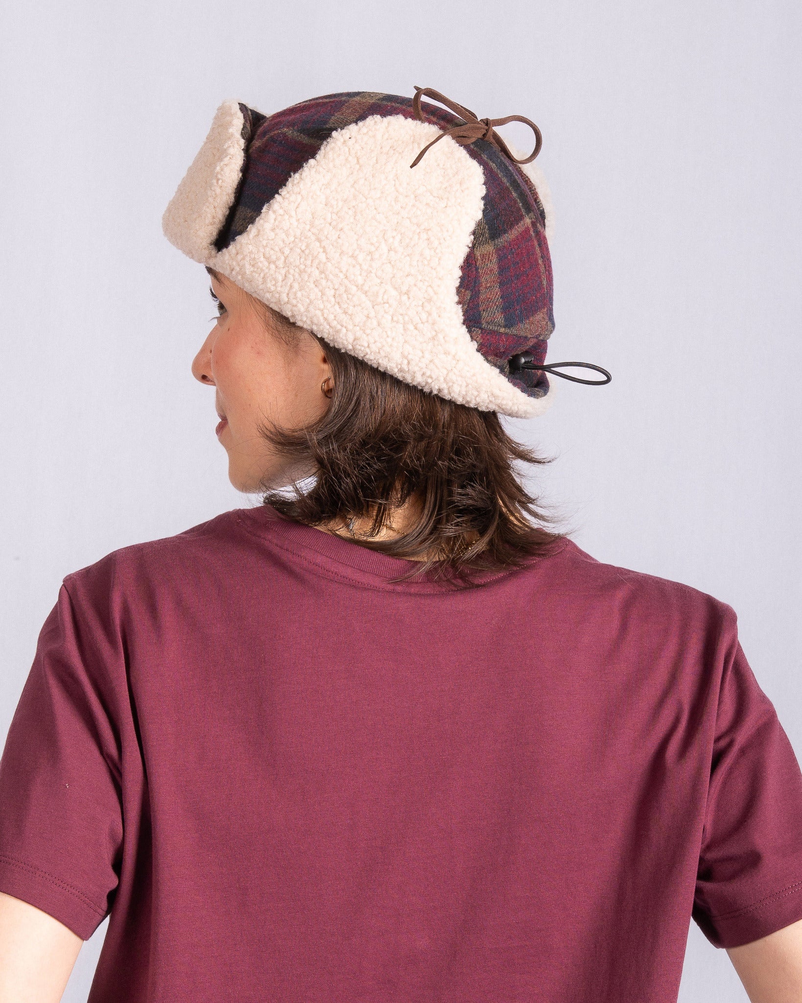 Person in a maroon shirt and plaid winter hat with shearling, shown from the back against a plain background.