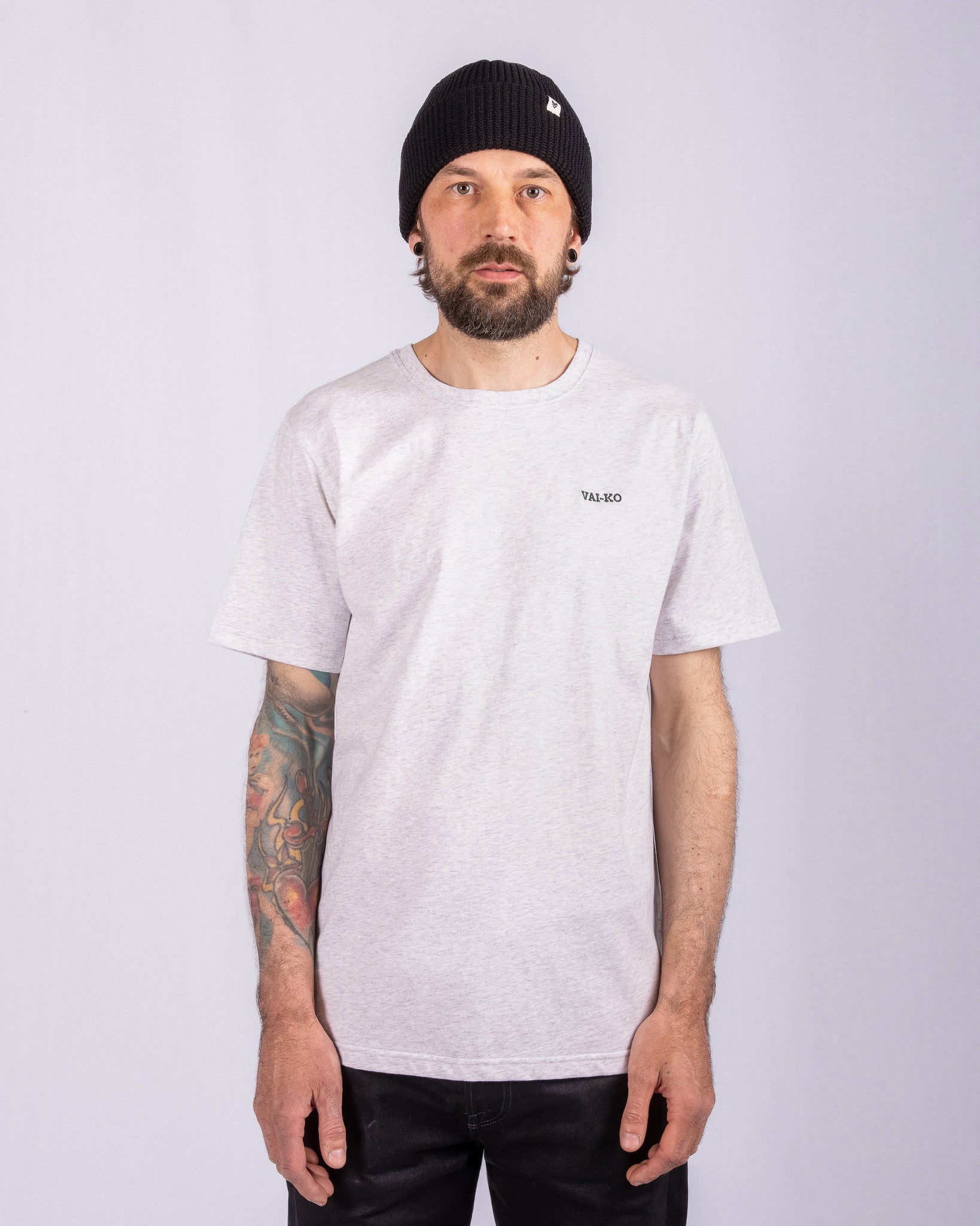 Man with a beard, wearing a black beanie and light gray t-shirt, stands against a plain background.