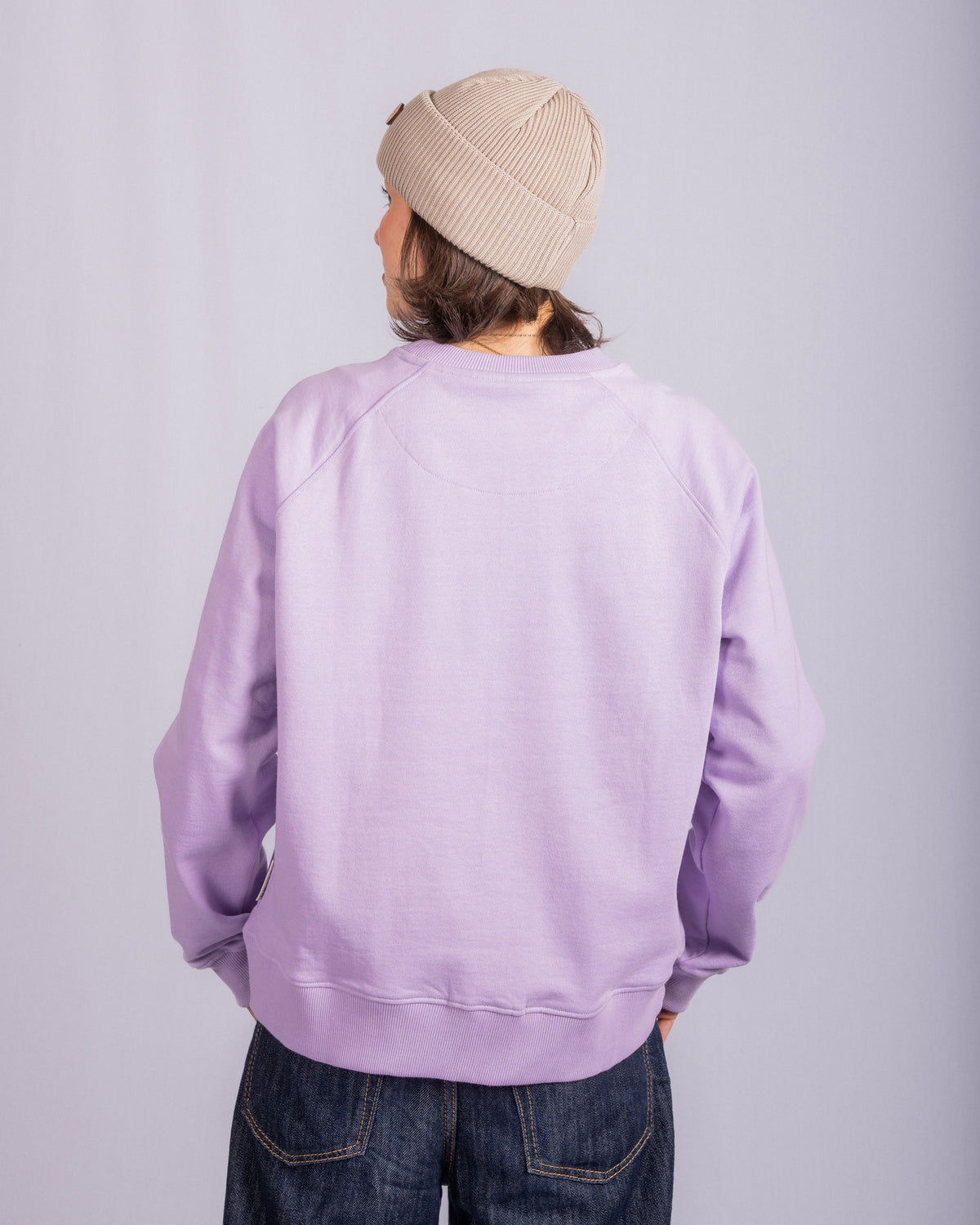 Person in a beige beanie and lilac sweatshirt stands facing away, hands on hips, against a plain background.