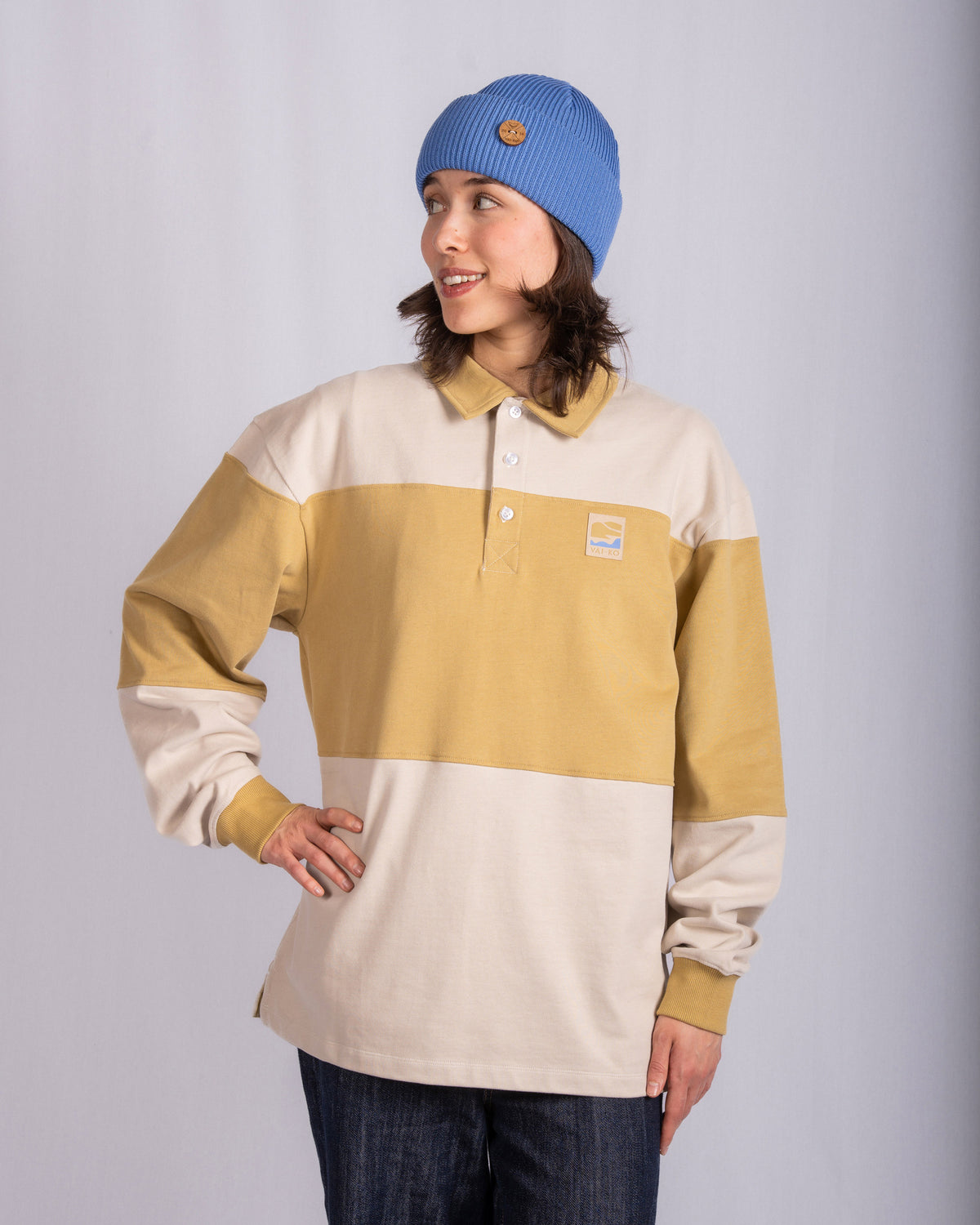 Person in blue beanie and beige-yellow striped shirt stands smiling, hand on hip, against plain backdrop.