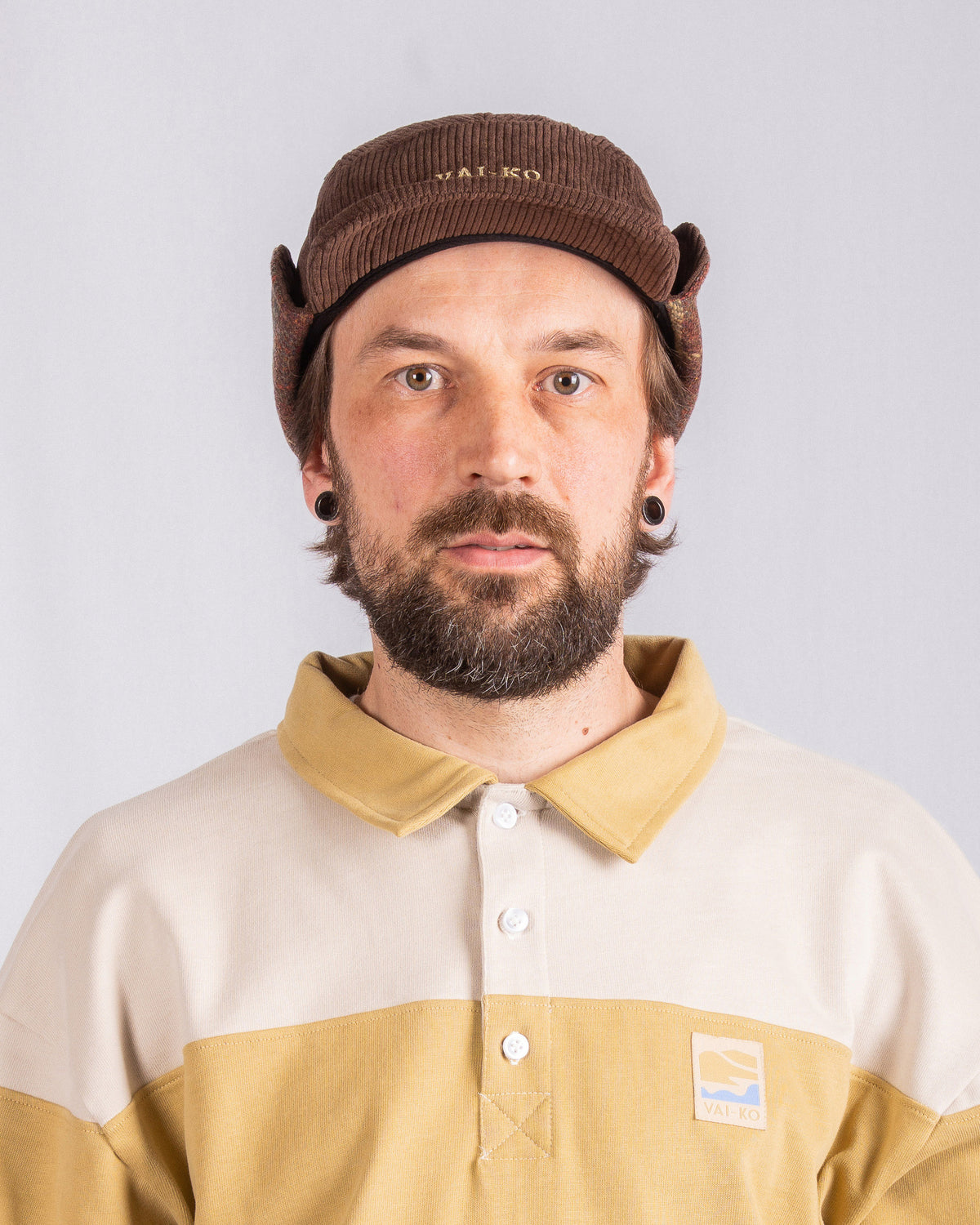 Man with beard wearing brown cap, cream and yellow shirt, looking at camera against plain background.
