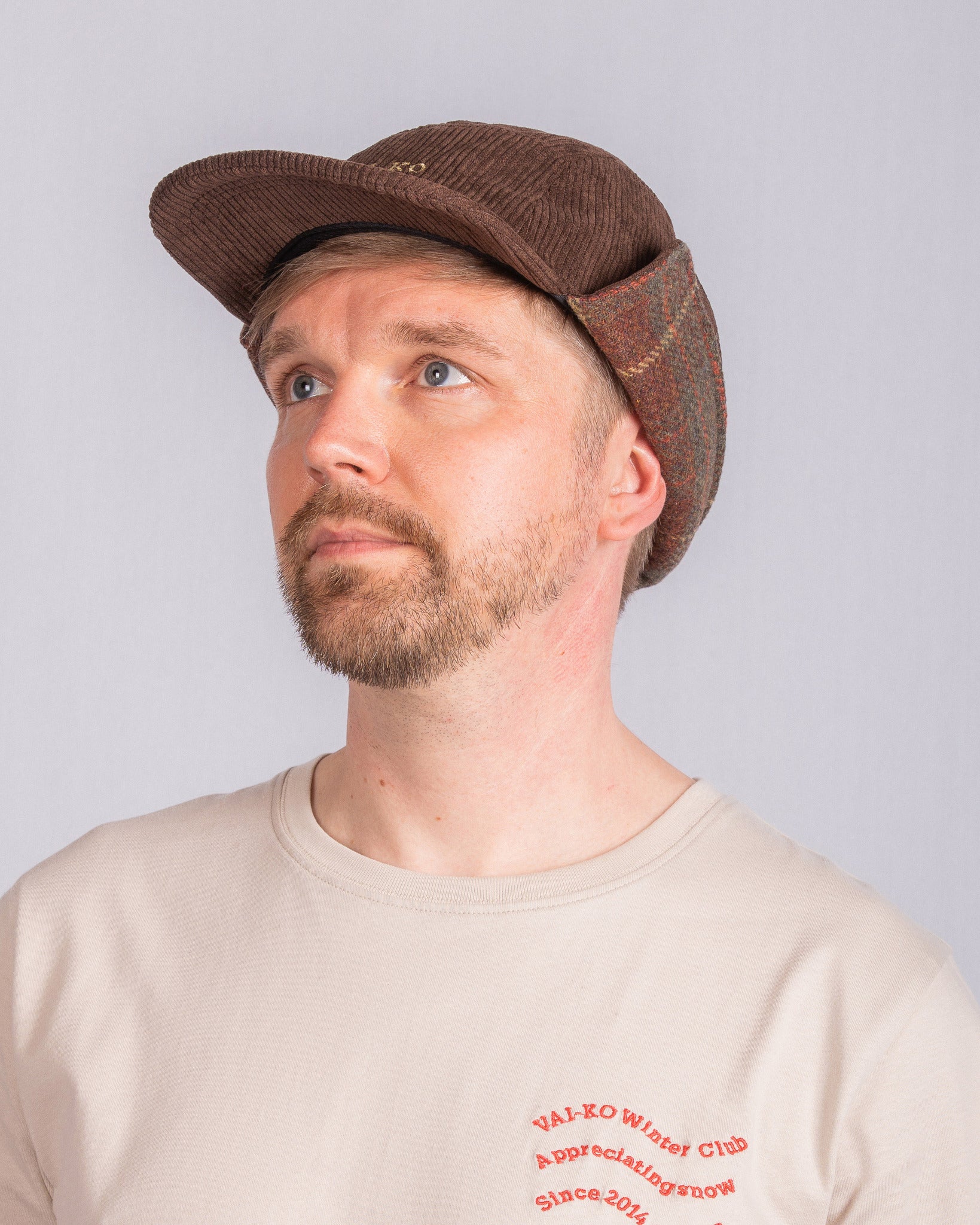 Man with a short beard wearing a brown cap and beige t-shirt, looking up against a plain background.