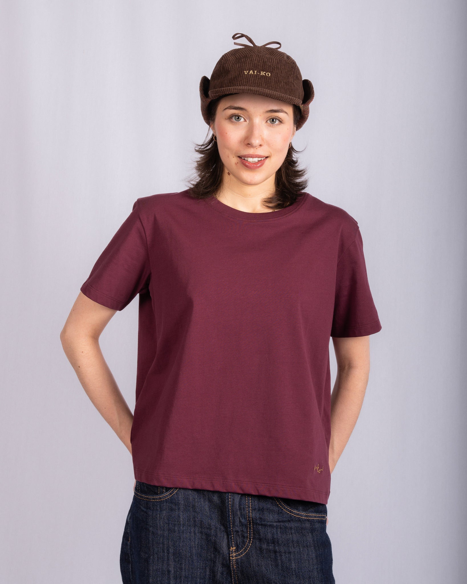 Person wearing a maroon t-shirt, dark jeans, and a brown corduroy hat, standing against a plain background.