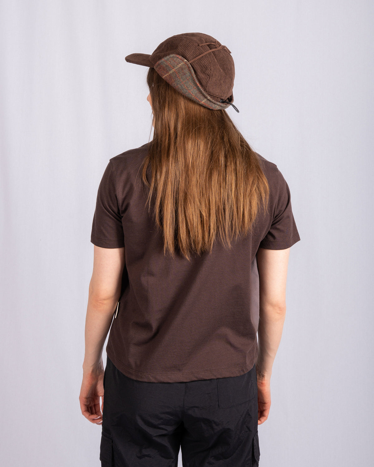 Person with long hair, brown t-shirt, and cap stands facing away from the camera against a plain background.