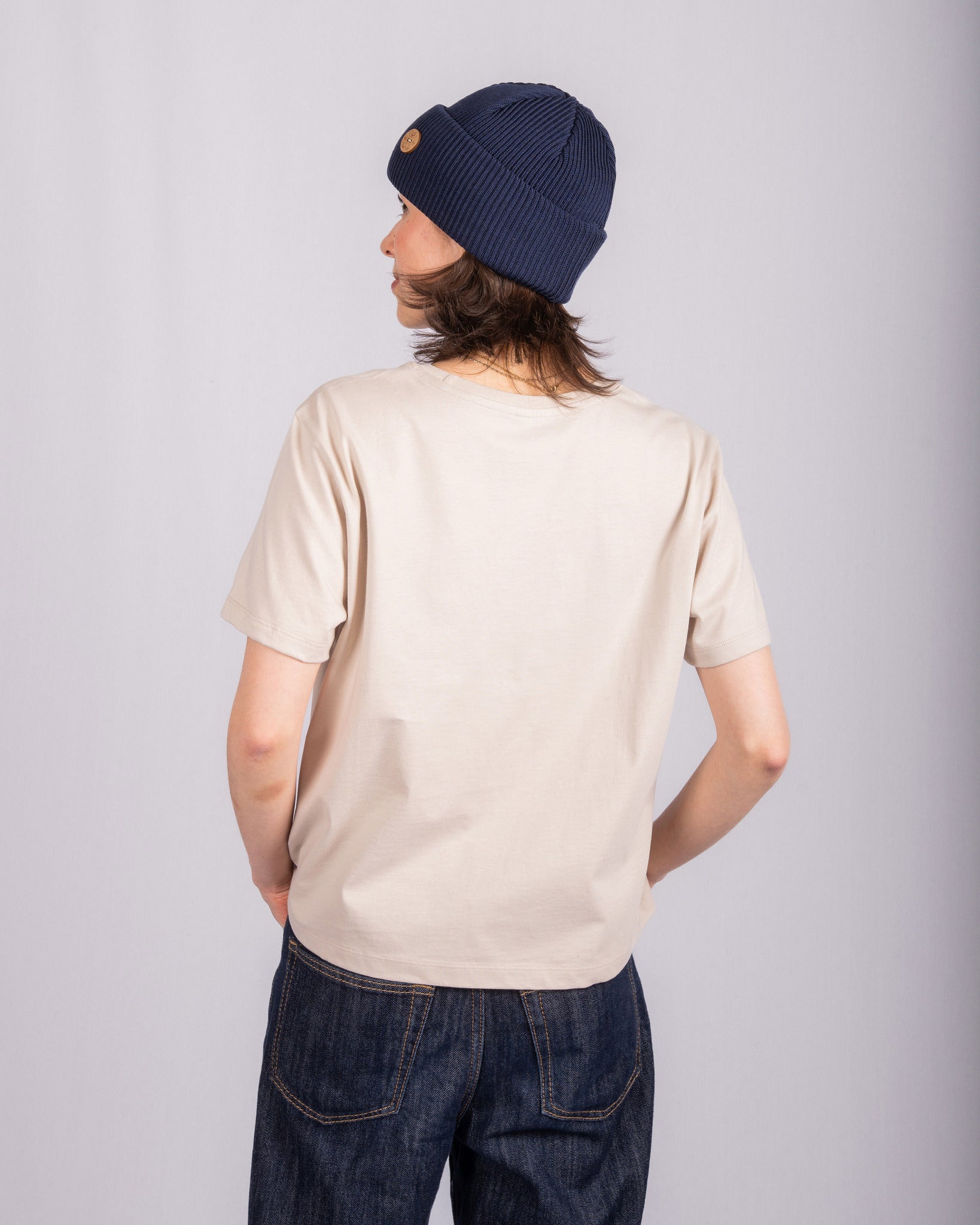 Person in a beige t-shirt and navy beanie stands with their back to the camera against a plain background.
