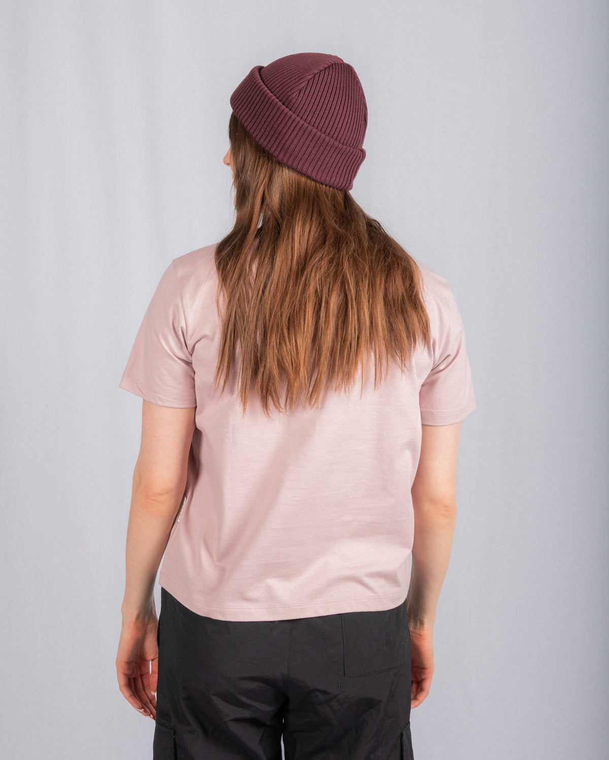Person in a mauve beanie and pink t-shirt faces away, standing against a plain light background.