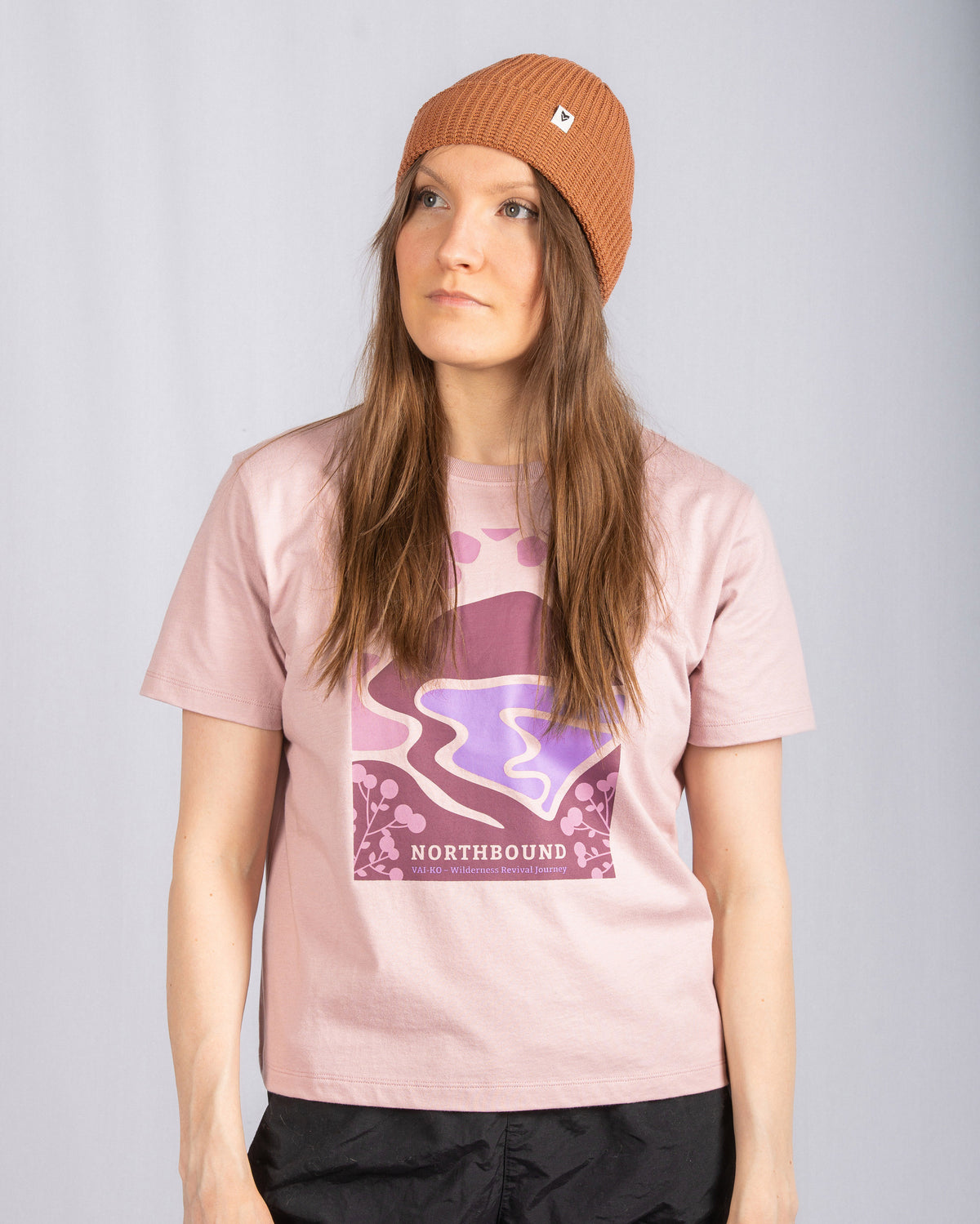 Woman in a brown beanie and pink graphic t-shirt stands against a plain light gray background.