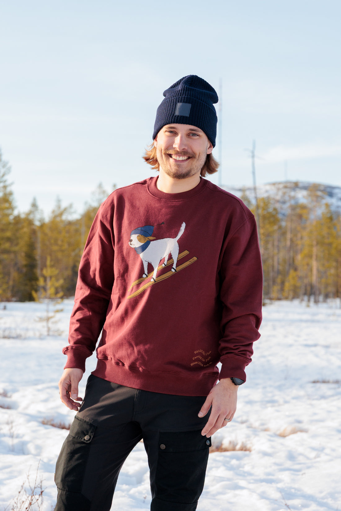 Smiling man in a maroon sweater with a dog print, standing on snow-covered ground with trees in the background.