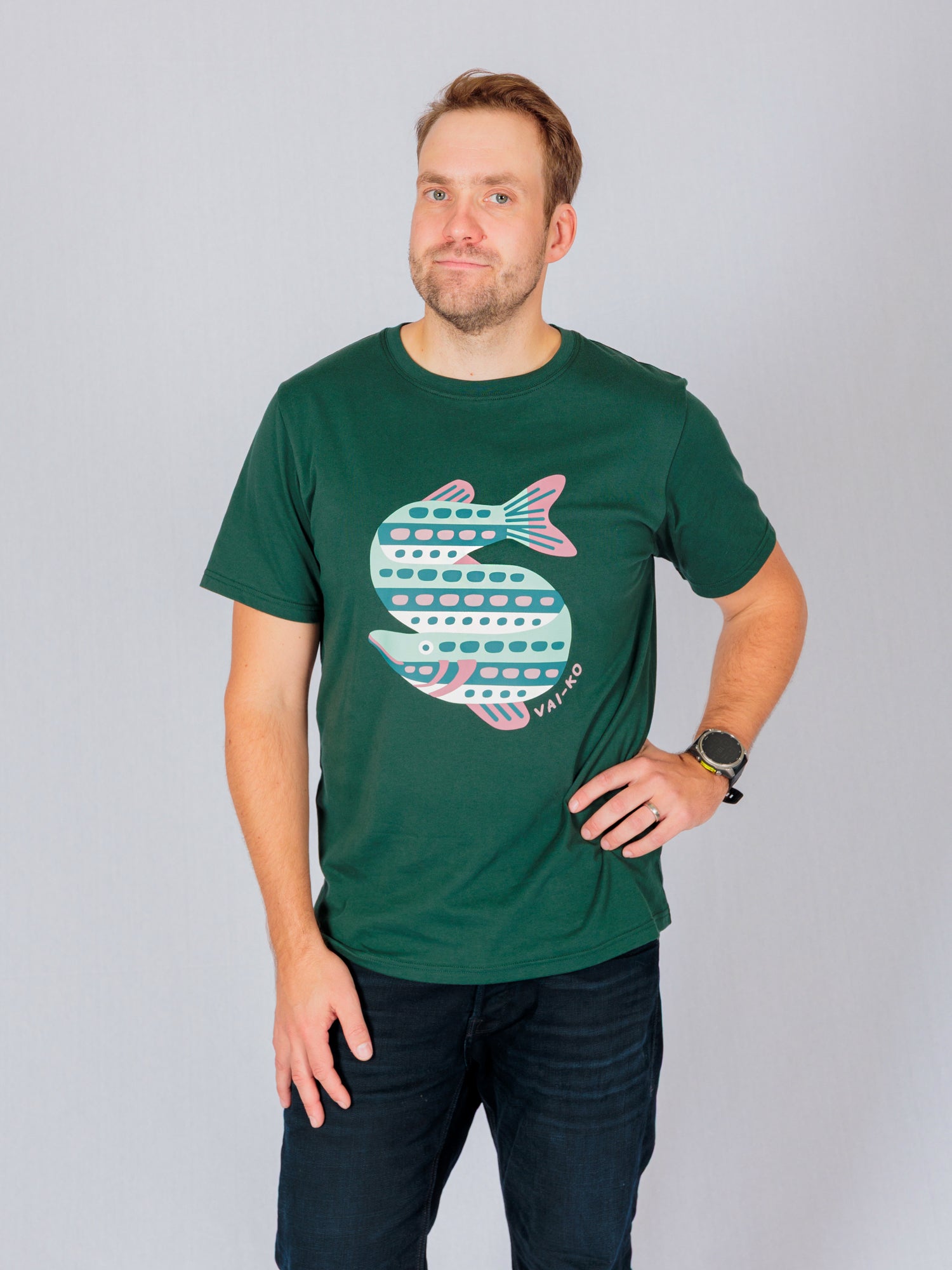 Man in a dark green t-shirt with a fish graphic, posing with one hand on his hip against a plain background.