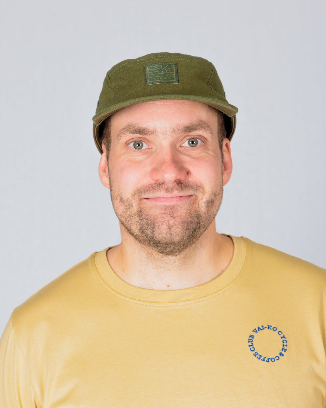 Smiling man in a yellow t-shirt and green cap standing against a plain, light gray background.
