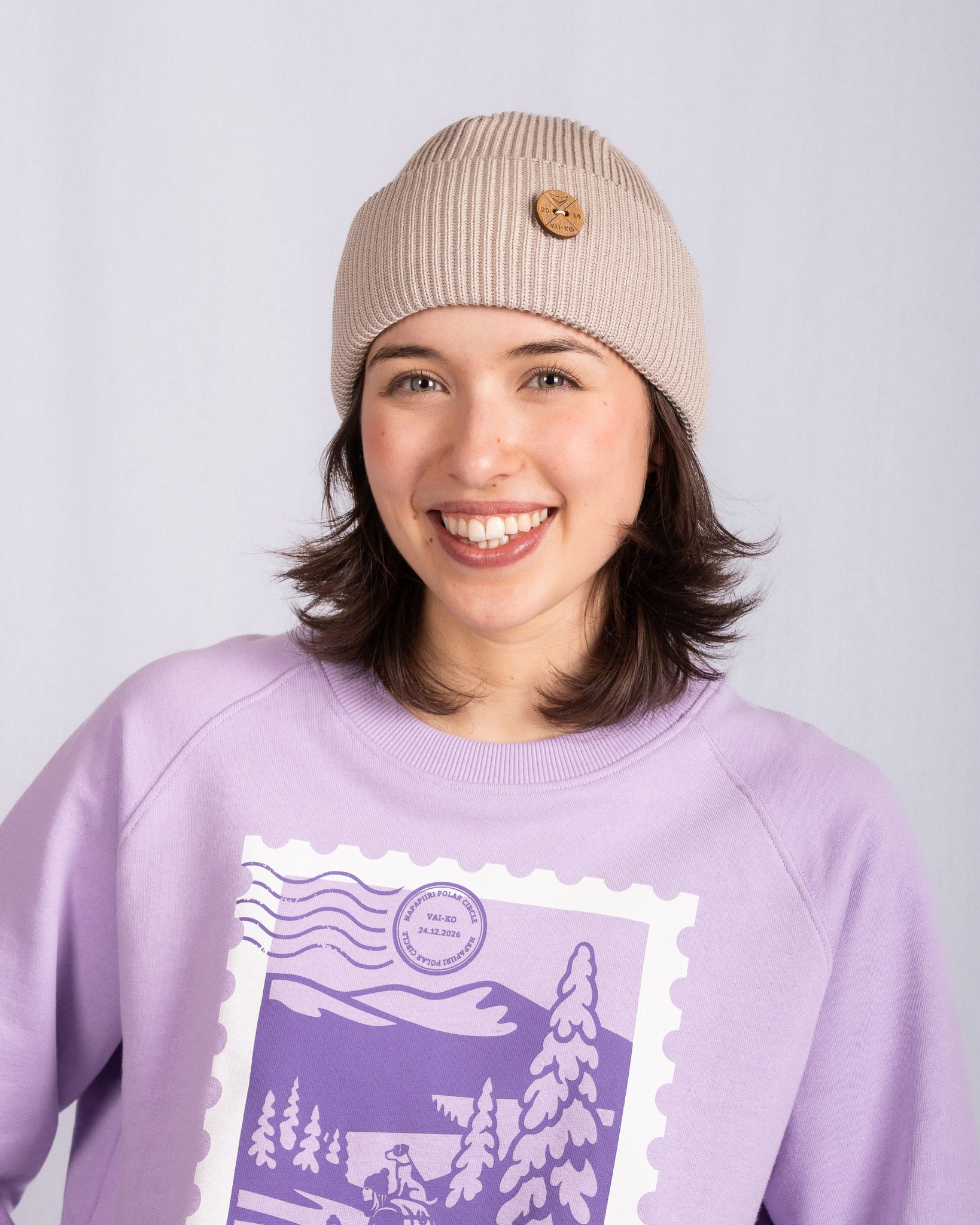 Smiling person in a beige beanie and lavender sweatshirt with a winter scene graphic, standing against a light background.