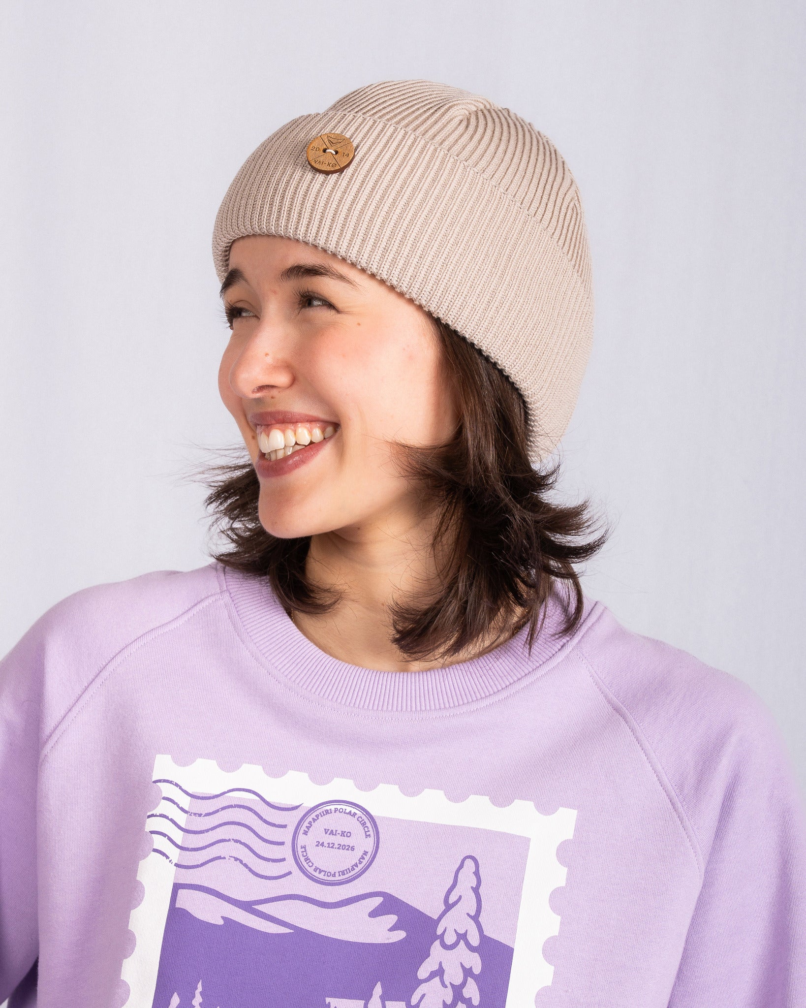 Smiling person in a beige beanie and lavender sweatshirt with a graphic, looking to the left.