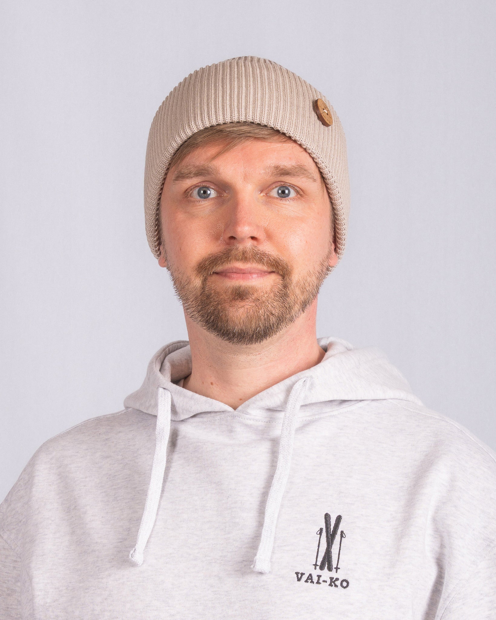 Man in beige knit beanie and light hoodie with VAI-KO logo, facing camera against plain background.