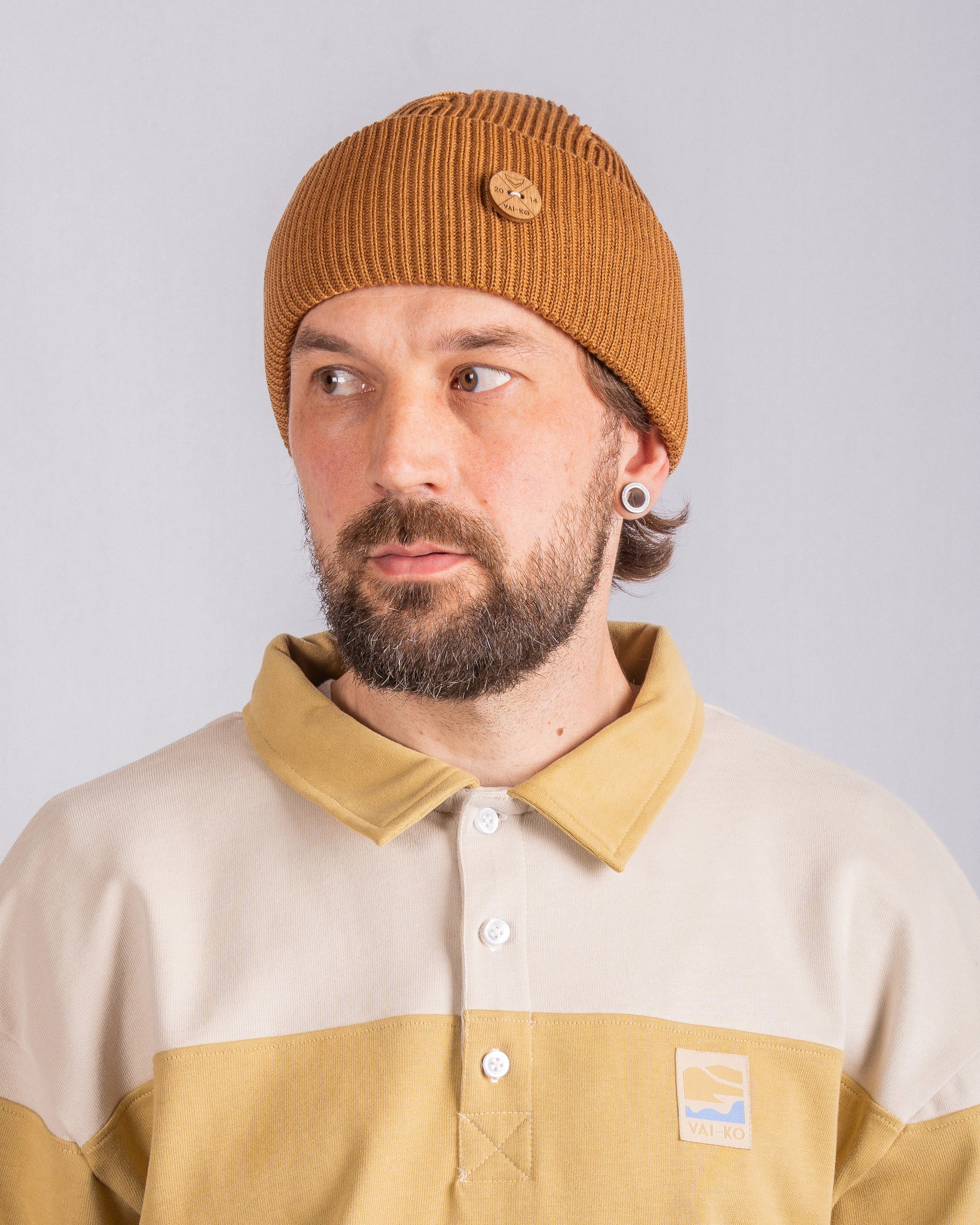 Man with brown beanie and beige shirt with logo, looking to his left against a plain background.