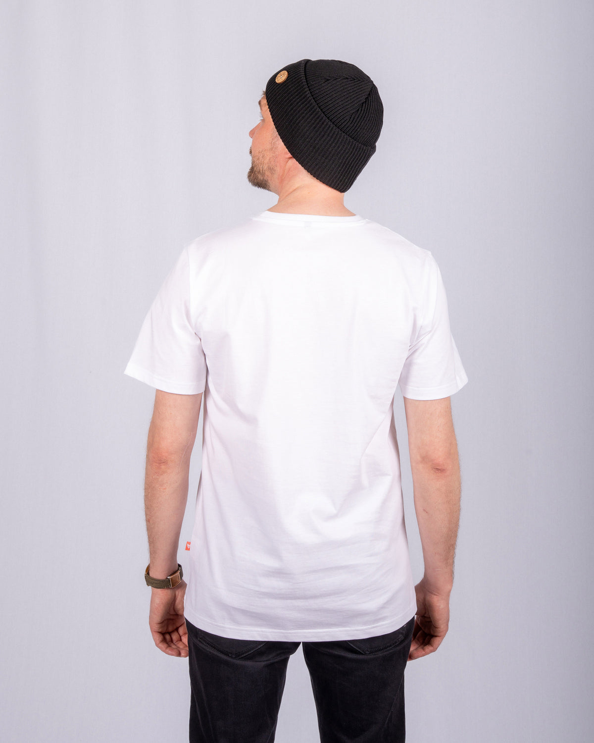 A man wearing a black beanie and white T-shirt stands facing away from the camera against a plain background.