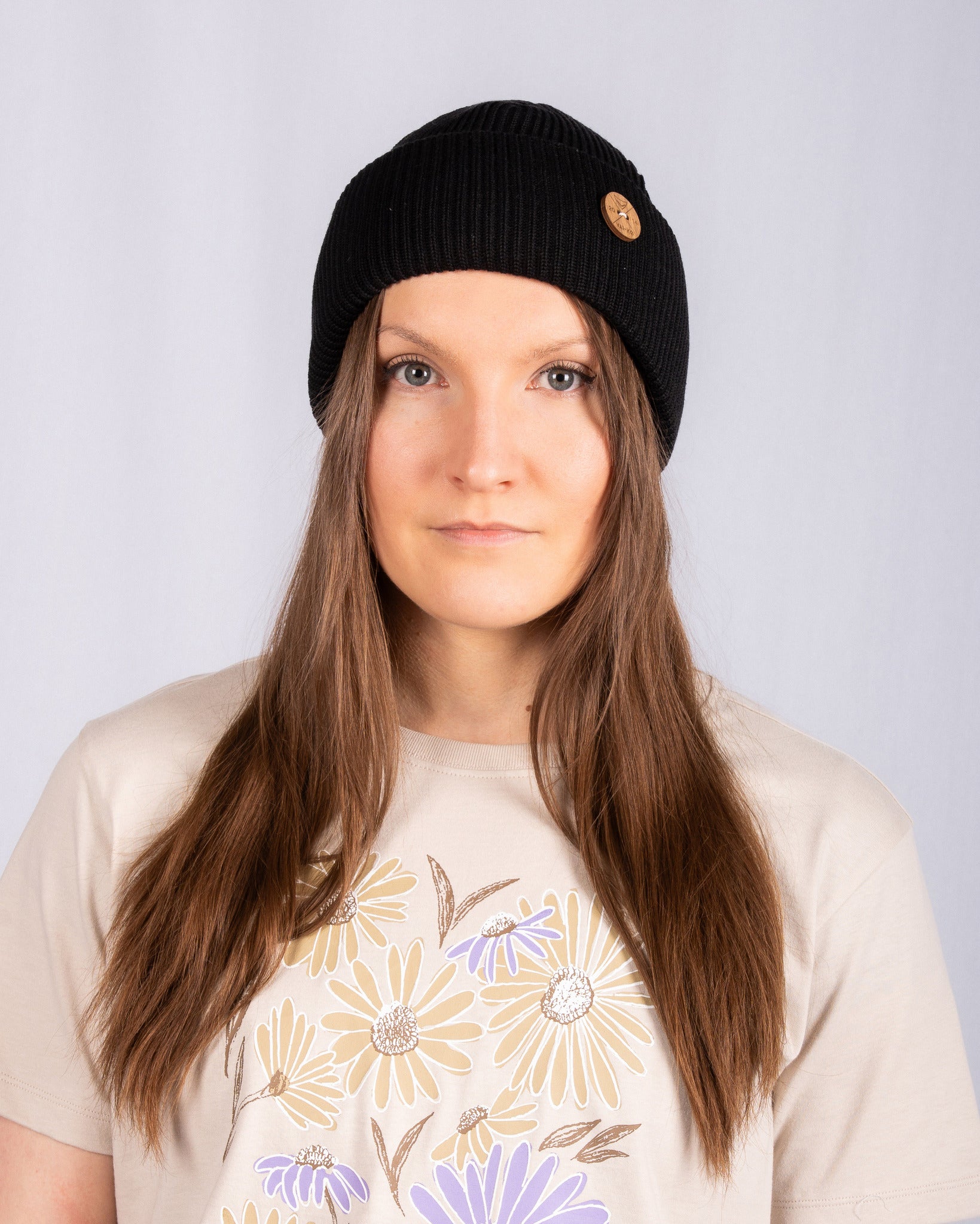 Woman with long brown hair wearing a black beanie and beige shirt with floral print, facing forward.