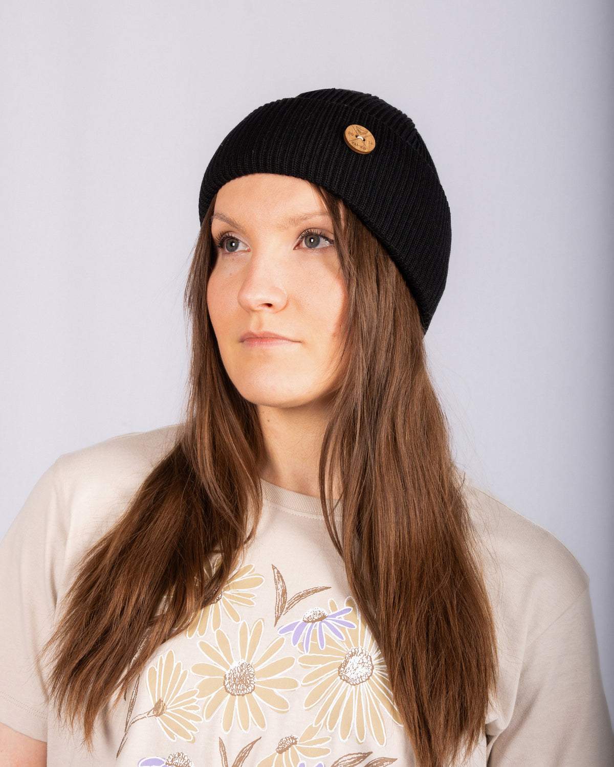 Woman with long brown hair wears a black beanie and beige floral shirt, looking slightly to the side.
